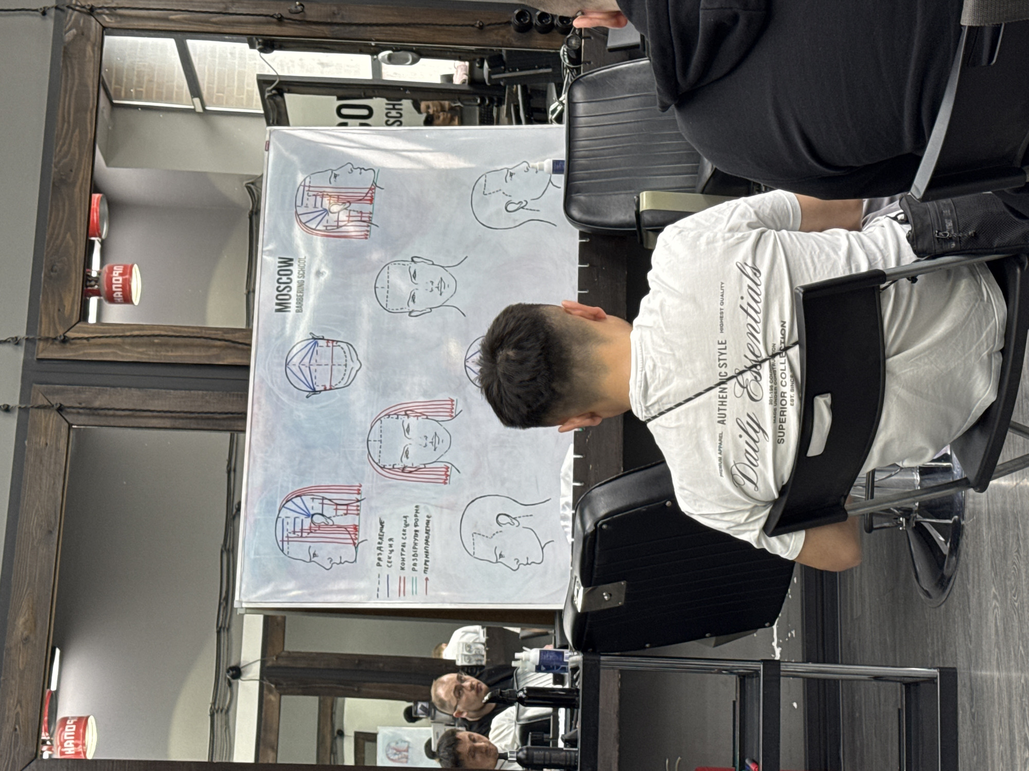 The image shows an indoor scene at the "MOSCOW BARBERING SCHOOL," likely a classroom or training area. In the foreground, a person with dark hair, wearing a white t-shirt, is seated in a black barber chair. They are positioned in front of a whiteboard displaying diagrams and text in what appears to be Russian, relating to haircutting techniques. Other individuals can be seen in the background reflected in a mirror. The room also contains barbershop-related furniture such as chairs and mirrors. The location is suggested by the name "MOSCOW BARBERING SCHOOL" on the whiteboard.