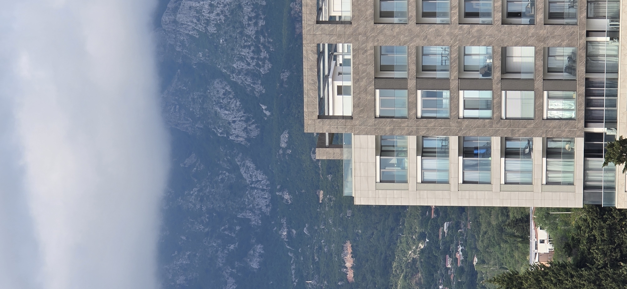 A low-angle, rotated view shows a modern multi-story building beside a mountainside in Bar, Montenegro. The building is tan-colored with a grid of rectangular windows. Some windows have blinds, others reflect the sky. To the left of the building is a forested mountain, partially obscured by white clouds. Small houses dot the landscape at the base of the mountain. The foliage indicates a natural setting near the city.