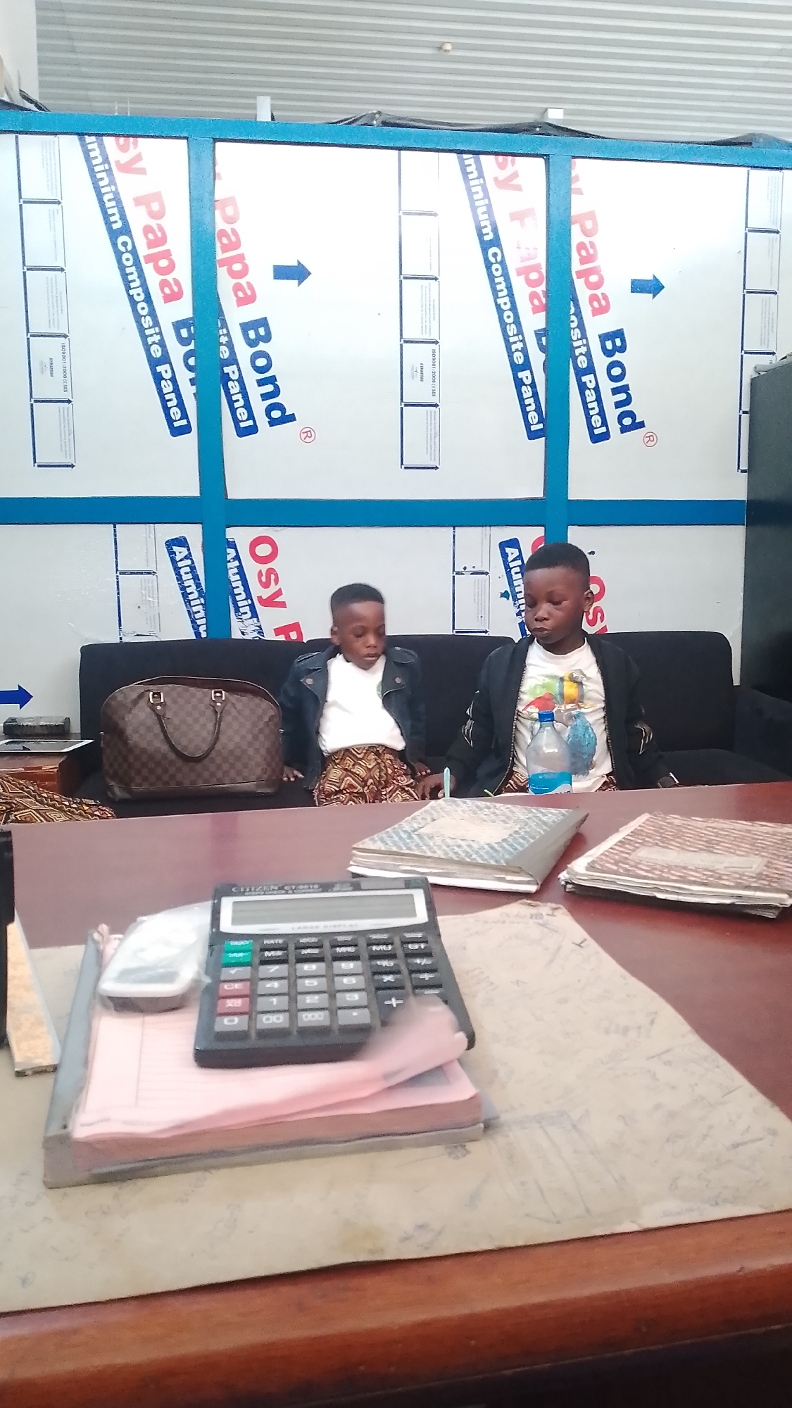 The image shows two young boys sitting on a black couch in front of a desk. They are both wearing jackets; the boy on the left has on a white shirt, while the boy on the right has on a graphic t-shirt. There is a brown Louis Vuitton bag to the left of the boy on the left, and a water bottle next to the boy on the right. On the desk are several books, a calculator, and some papers. The background features panels with the words "Papa Bond" and "Aluminium Composite Panel". The location of this photo is in Orerokpe, Nigeria.