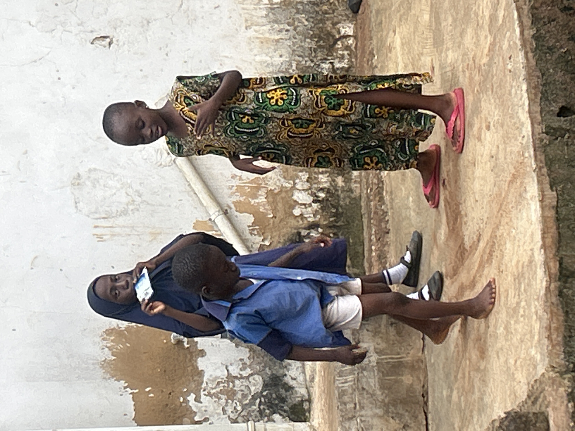 Three children, two girls and a boy, stand on a concrete surface in front of a weathered, textured wall. The girl on the left wears a dark blue hijab and a long dark blue garment, and holds a small, rectangular object, possibly a card or packet, to her mouth. The boy in the center wears a light blue short-sleeved collared shirt, white shorts, and dark shoes with white socks. He is looking down. The girl on the right wears a colorful, patterned dress with a slit up the side, and pink flip-flops. Her left hand is raised to her chest. The environment appears to be an outdoor urban setting in Ikorodu, Nigeria, characterized by the rough texture of the wall and the concrete ground, with some debris visible in the lower foreground.