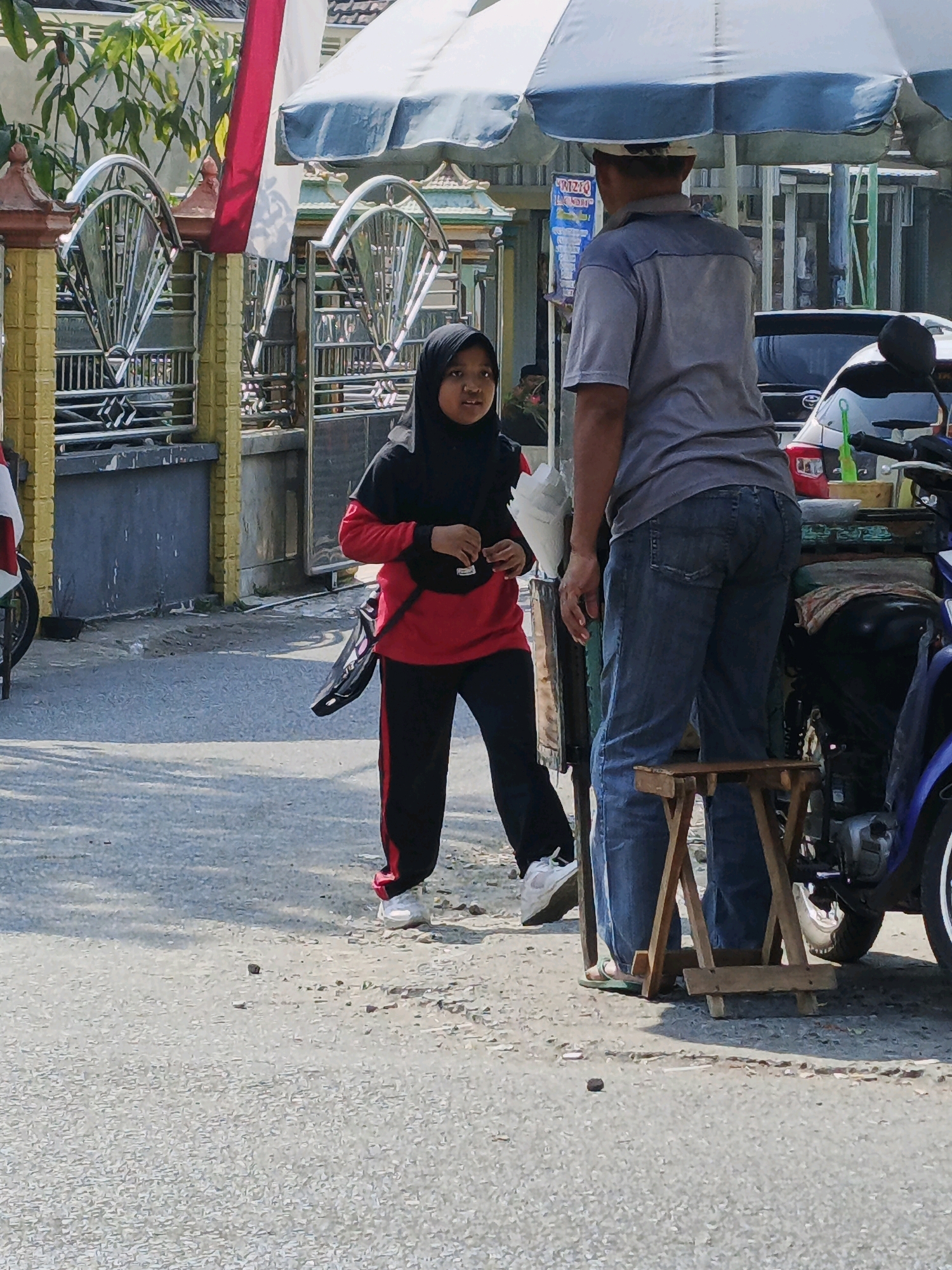 In Ketegan, Indonesia, a young girl wearing a black hijab and a red and black tracksuit stands on a paved street, facing a man. The girl is looking towards the man, who is wearing a gray t-shirt and blue jeans, and is standing next to a cart with an umbrella overhead. The cart is laden with various items, including what appears to be food or drinks, and is attached to a motorcycle on the right side of the frame. Behind the girl, a yellow and silver gate with intricate metalwork is visible, as well as a white and red Indonesian flag. The street is littered with small stones and debris, and shadows indicate it is daytime. The scene suggests a street vendor interaction or a casual encounter between two individuals.