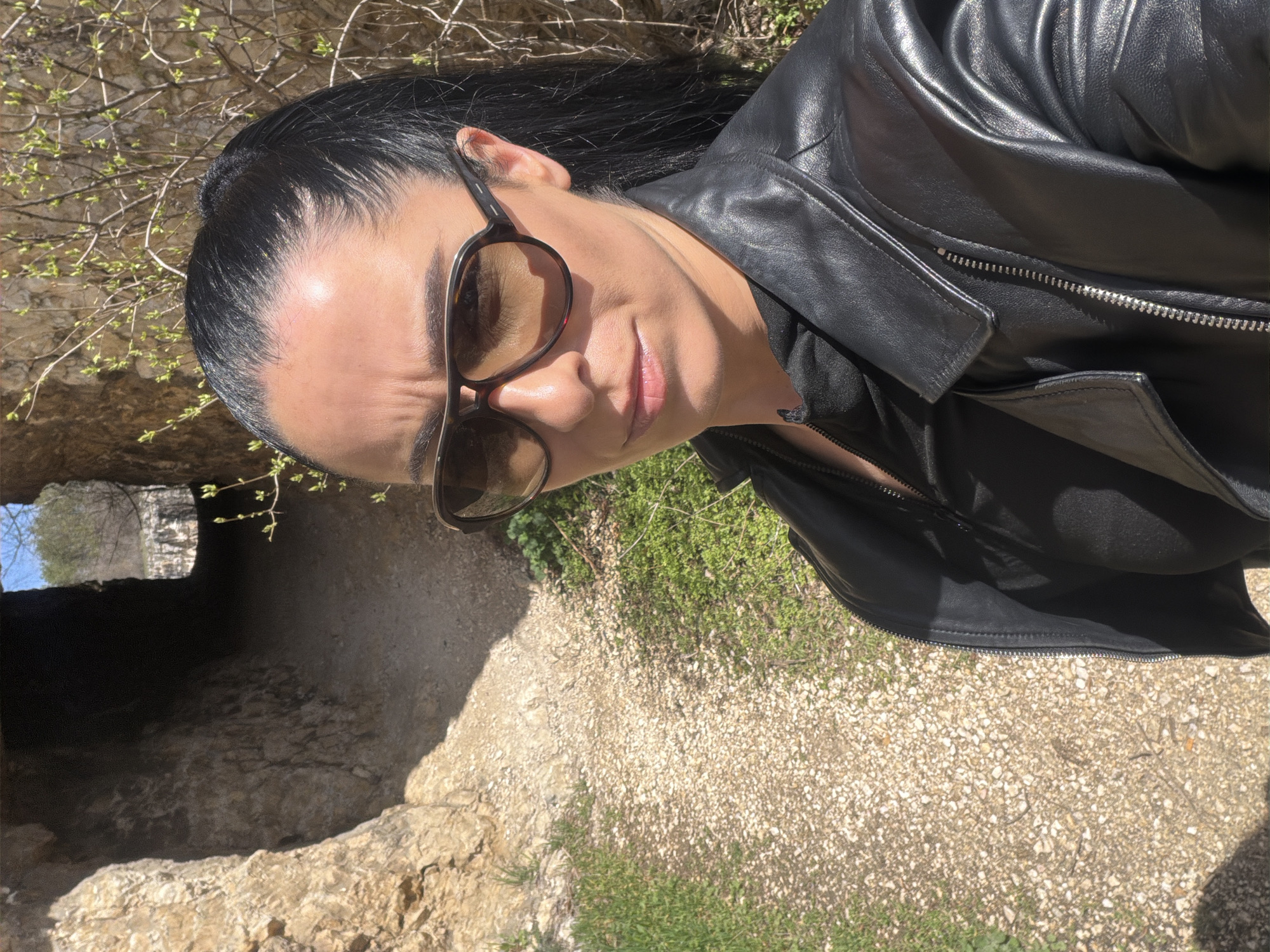 A female individual is depicted in a medium-shot selfie, standing on an unpaved, gravel path within a rocky passage, identified as being in Kaylaka Park, Pleven, Bulgaria. She has dark hair styled in a high ponytail and is wearing dark aviator-style sunglasses with brown lenses and a dark, possibly tortoise-shell, frame. Her expression is neutral as she directly faces the camera. She is dressed in a black leather jacket featuring a prominent silver zipper and an open collar, worn over a dark, possibly black, inner garment.
To the subject's left, a light brown, rough-hewn rock face forms the wall of the passage. The path, comprised of light-colored gravel and soil, extends behind her and narrows into a dark, tunnel-like opening cut into the rock. A distant, bright light source is visible at the far end of this tunnel. On both sides of the path, particularly to the subject's right, green grass and early spring foliage, characterized by bare branches with small, light green leaves, are present. The scene is illuminated by strong, natural sunlight, casting visible highlights and shadows.