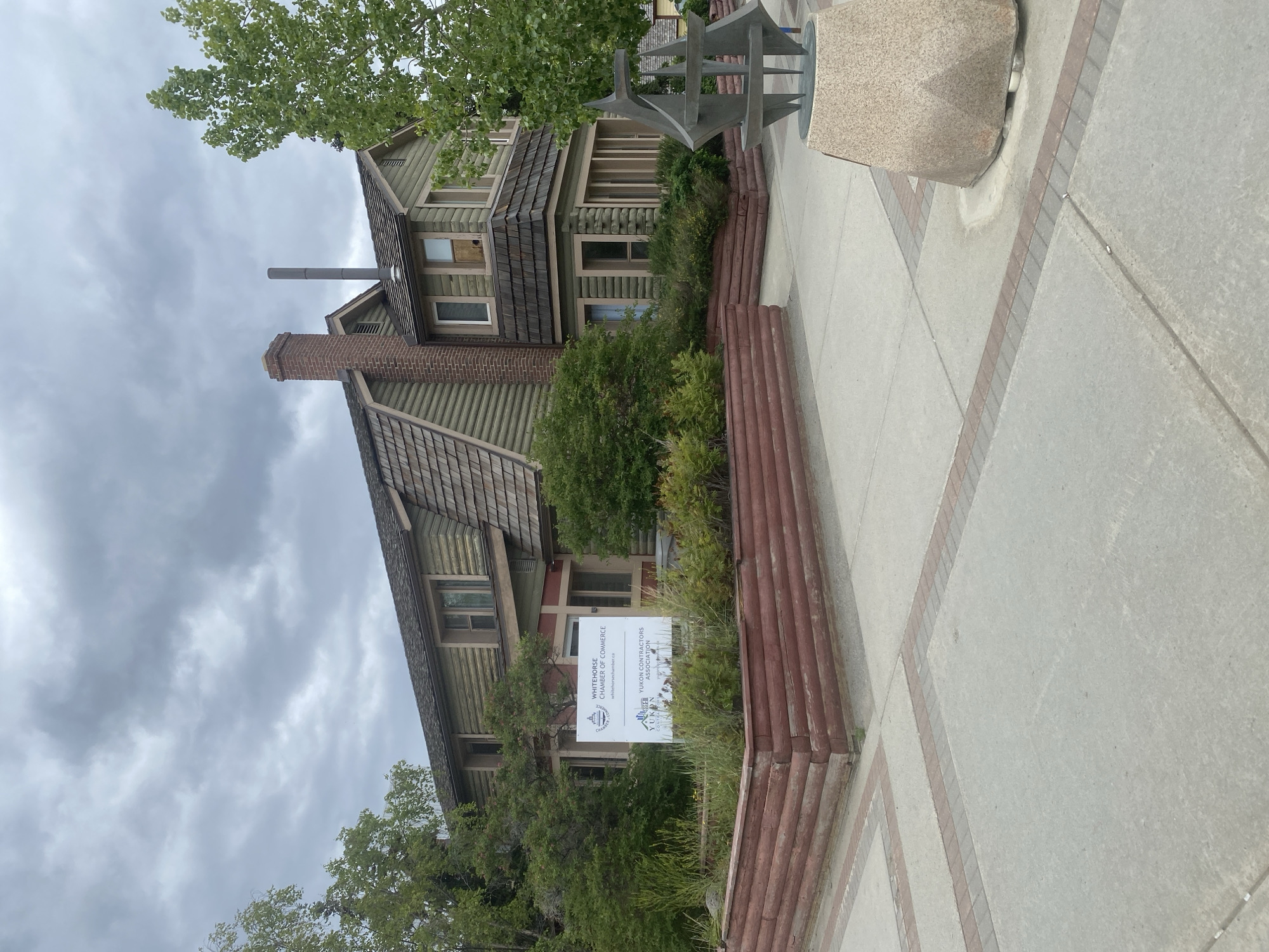 The image depicts the Whitehorse Chamber of Commerce building located in Whitehorse, Yukon, Canada. The building is a two-story structure with a rustic design, featuring wooden siding and a shake roof. A prominent brick chimney rises from the roof. In front of the building, a sign displays "WHITEHORSE CHAMBER OF COMMERCE" and "YUKON CONTRACTORS ASSOCIATION." A wooden planter box filled with greenery is situated between the building and the paved walkway. A modern metal sculpture rests on a stone pedestal to the right of the walkway. The sky is overcast with clouds, and trees surround the building. The paved walkway is constructed with concrete slabs and bordered by brick accents.
