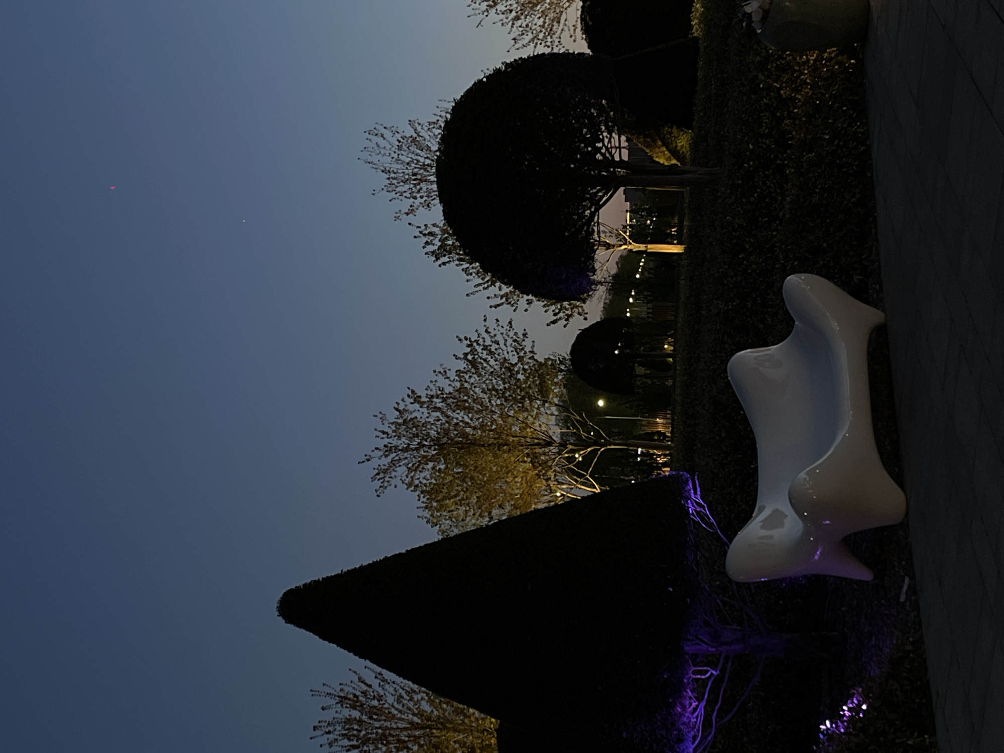A nighttime outdoor scene shows a modern white bench next to dark bushes and trees against a dark blue sky. In the distance, lights suggest a city setting. The location is possibly Krasnodar, Russia.