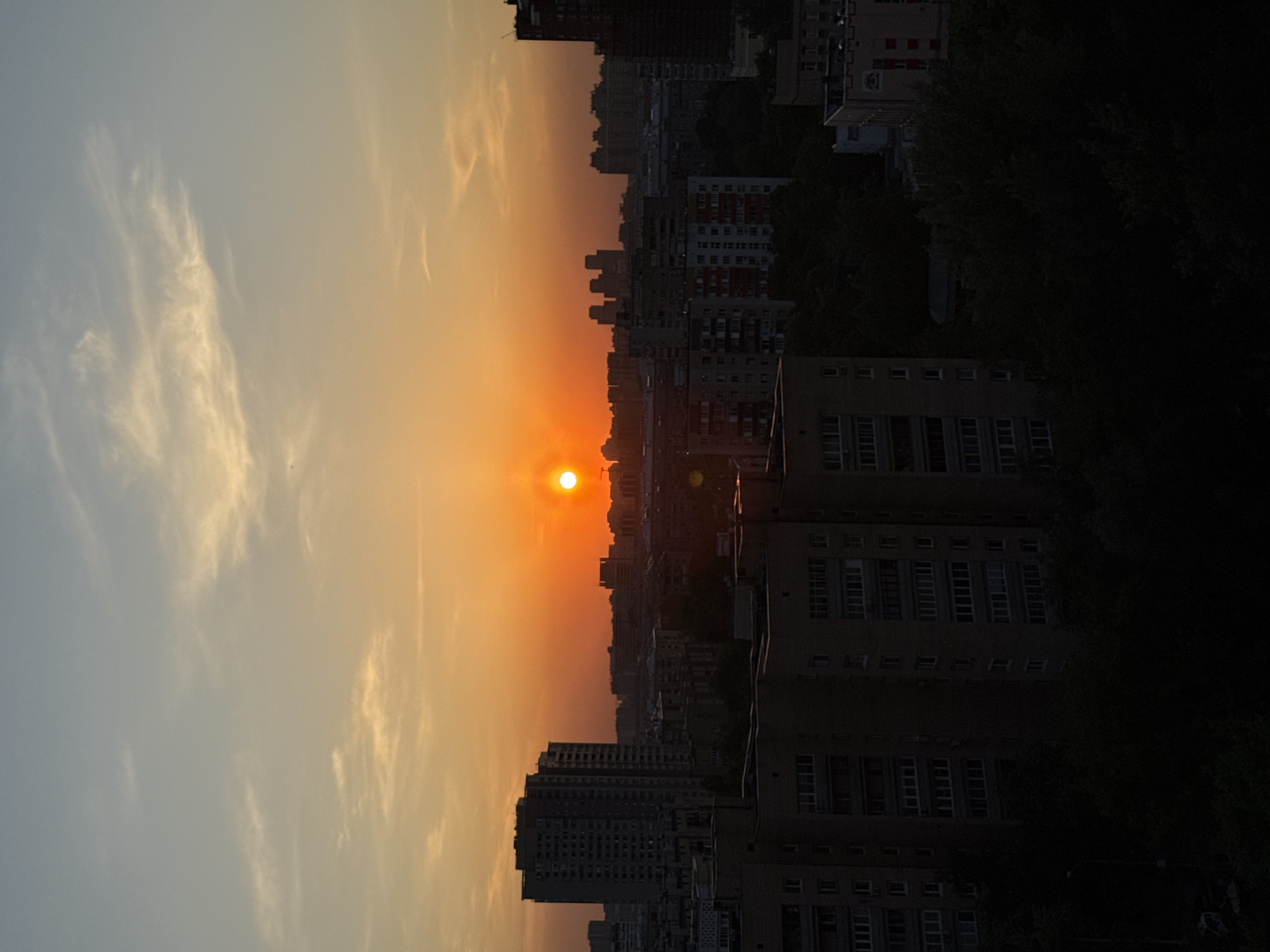 The image captures a sunset over an urban skyline in Moscow, Russia. The sun, a bright orange orb, is positioned near the horizon, casting an orange and yellow glow across the sky. The sky on the left side of the image shifts to a paler blue, with scattered white clouds. The silhouette of multiple buildings forms the skyline, ranging from apartment blocks to taller high-rises. Some trees appear dark along the right edge of the image, adding a natural element to the cityscape. The overall scene conveys a peaceful, urban sunset.
