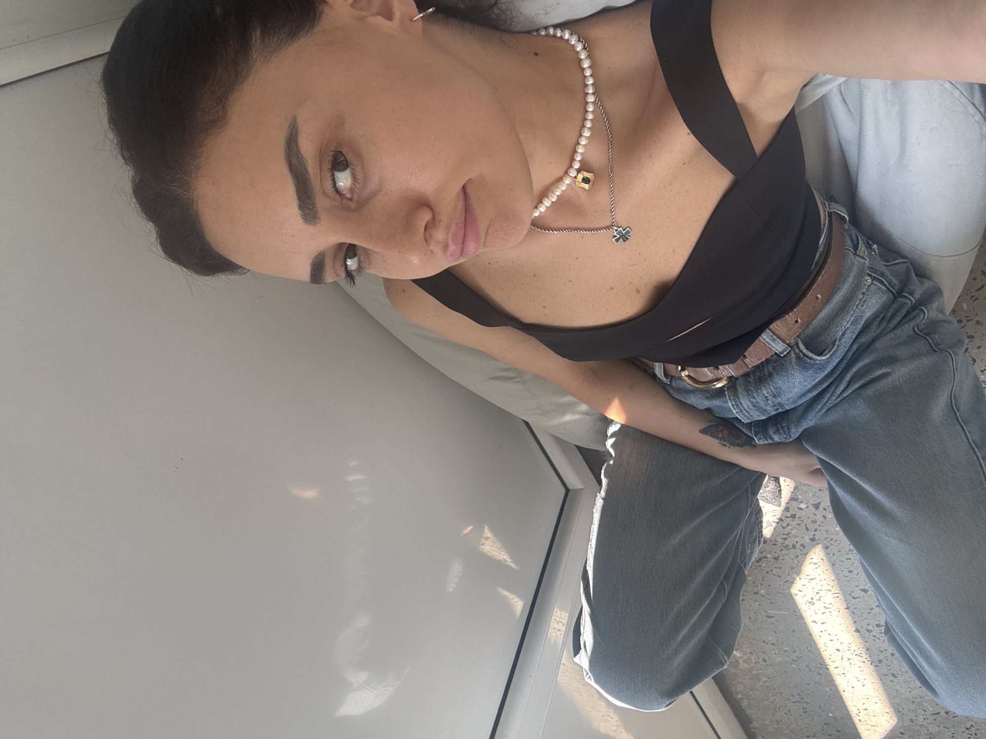 The image shows a woman in a black top and jeans, lying down. She is wearing a pearl necklace with a pendant. The environment appears to be indoors, with a white ceiling and a gray wall. Sunlight is casting shadows on the floor, suggesting it is daytime. The woman's legs are crossed and she is looking towards the camera. There is a tattoo of a butterfly on her left arm. The overall impression is of a casual and relaxed setting. The specific location in Tbilisi, Georgia, cannot be determined from the provided image.