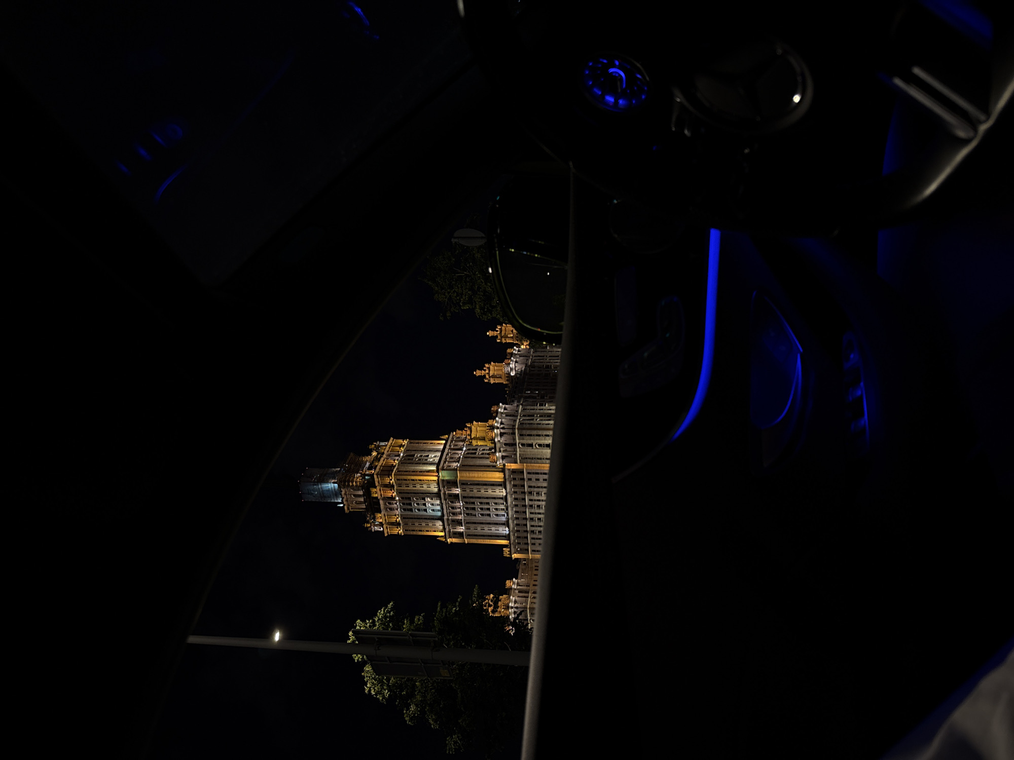 The image, oriented incorrectly and appearing rotated 90 degrees clockwise, captures a nighttime view from inside a vehicle. The primary subject is a large, ornate building, characteristic of Stalinist architecture, illuminated with warm yellow lights against a dark sky. The building features multiple stories, numerous windows, and intricate architectural details, suggesting a significant landmark in Moscow. Below the building, parts of illuminated trees and a street light are visible.
The scene is framed by elements of the car's interior. On the top portion of the image (when correctly oriented), blue ambient lighting is visible on ceiling controls, a circular dial, and along a side panel, suggesting a modern car interior. No specific activity beyond observing the illuminated architecture from the car is evident. The time of day is clearly night, with artificial lights defining the scene. There are no people visible, and no discernible text appears in the image.