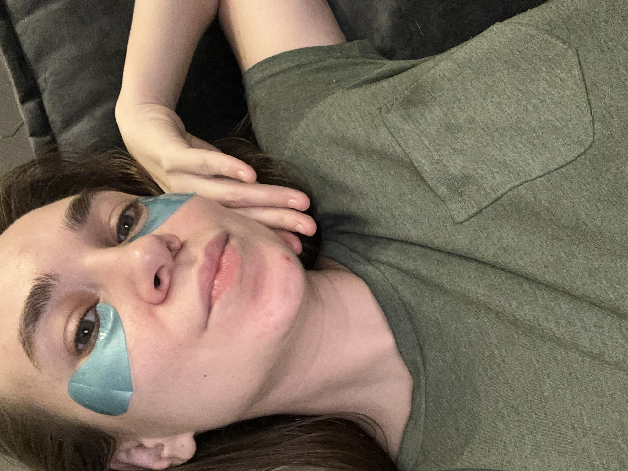 A woman is lying on a gray couch wearing an olive green t-shirt. She has eye masks on. The location is unknown, and no evidence indicates the city of Kharnavy, Russia.