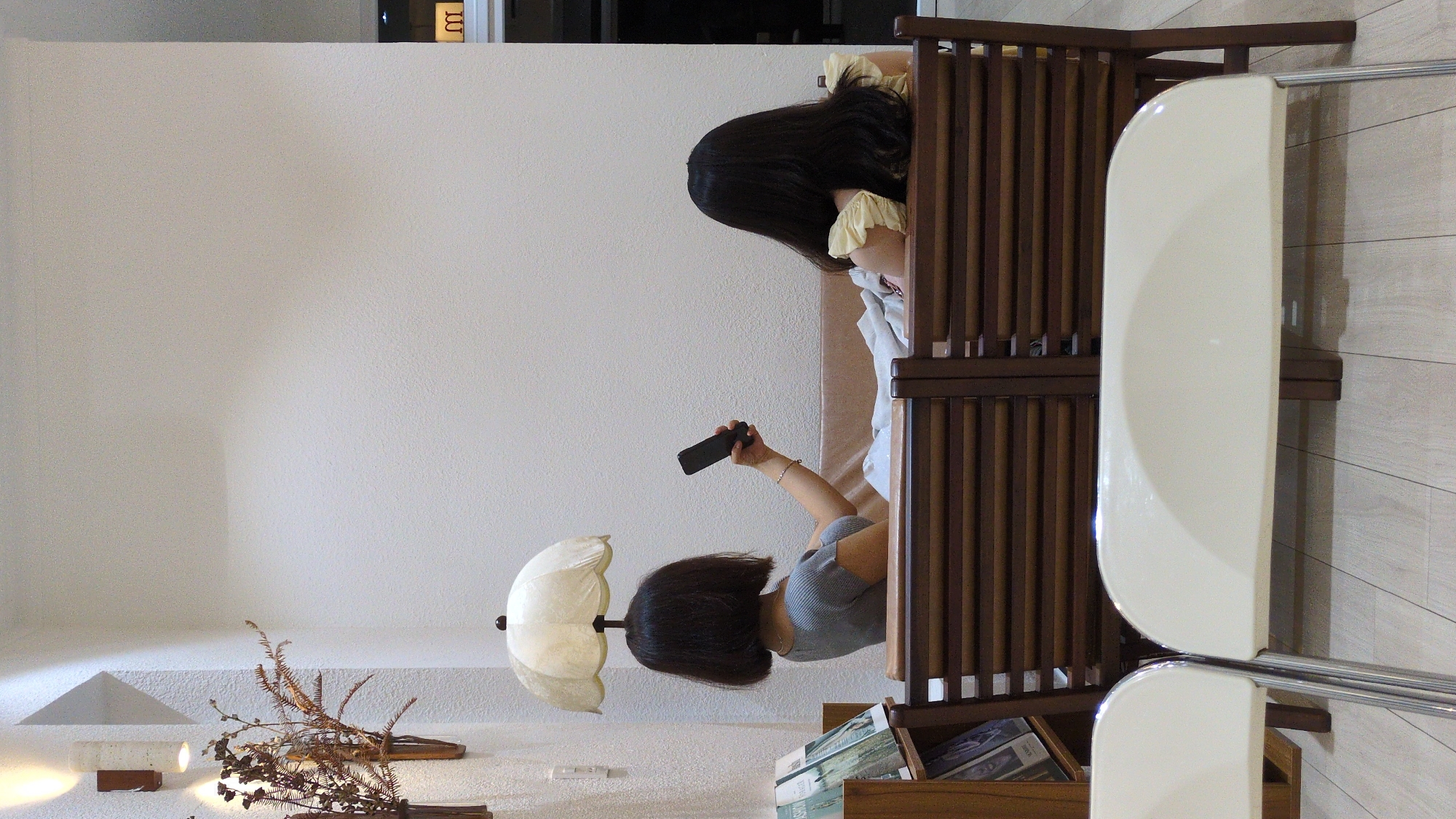 Two women are in a room. One woman is lying on a bed inside a wooden partition. She is looking at a cell phone in her hand. The other woman stands near an umbrella lamp on a table. The room is likely located in Phan Rang - Tháp Chàm, Vietnam. A white wall is visible, and there are decorative plants on a shelf. A white chair with a metal frame sits to the right.