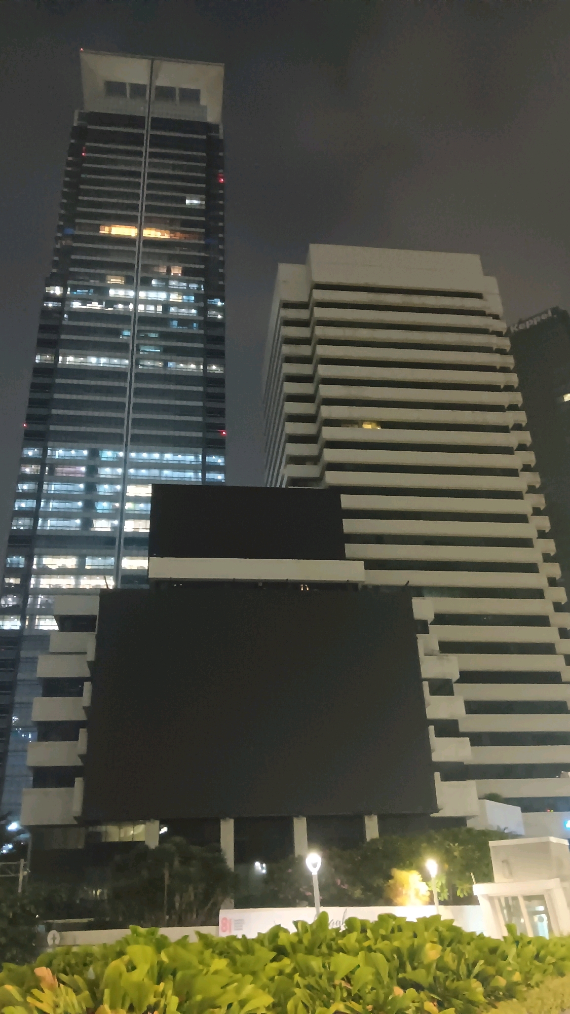 This is a nighttime, low-angle, outdoor, vertical photograph of a cluster of modern buildings in South Jakarta, Indonesia. The primary focus is on two tall skyscrapers and a large, dark, rectangular LED billboard.
The skyscraper on the left is dark with numerous floors, each featuring a grid of windows, many of which are illuminated with warm yellow and white light, suggesting interior illumination. The building has a distinctive white, tiered roof structure.
To the right of the left skyscraper is a wider, white building with a stepped facade composed of many horizontal bands, creating a textured appearance. Some of the windows in this building are also lit. Partially visible on the far right, a section of another dark building is seen, with the word "Keppel" faintly discernible on it.
Dominating the foreground and middle ground is a very large, dark, rectangular LED billboard. It appears to be mounted on a lower section of the white stepped building. Below the billboard, there is a landscaped area with lush green foliage. Two streetlights with a warm glow are visible amongst the plants. The sky is dark and overcast.