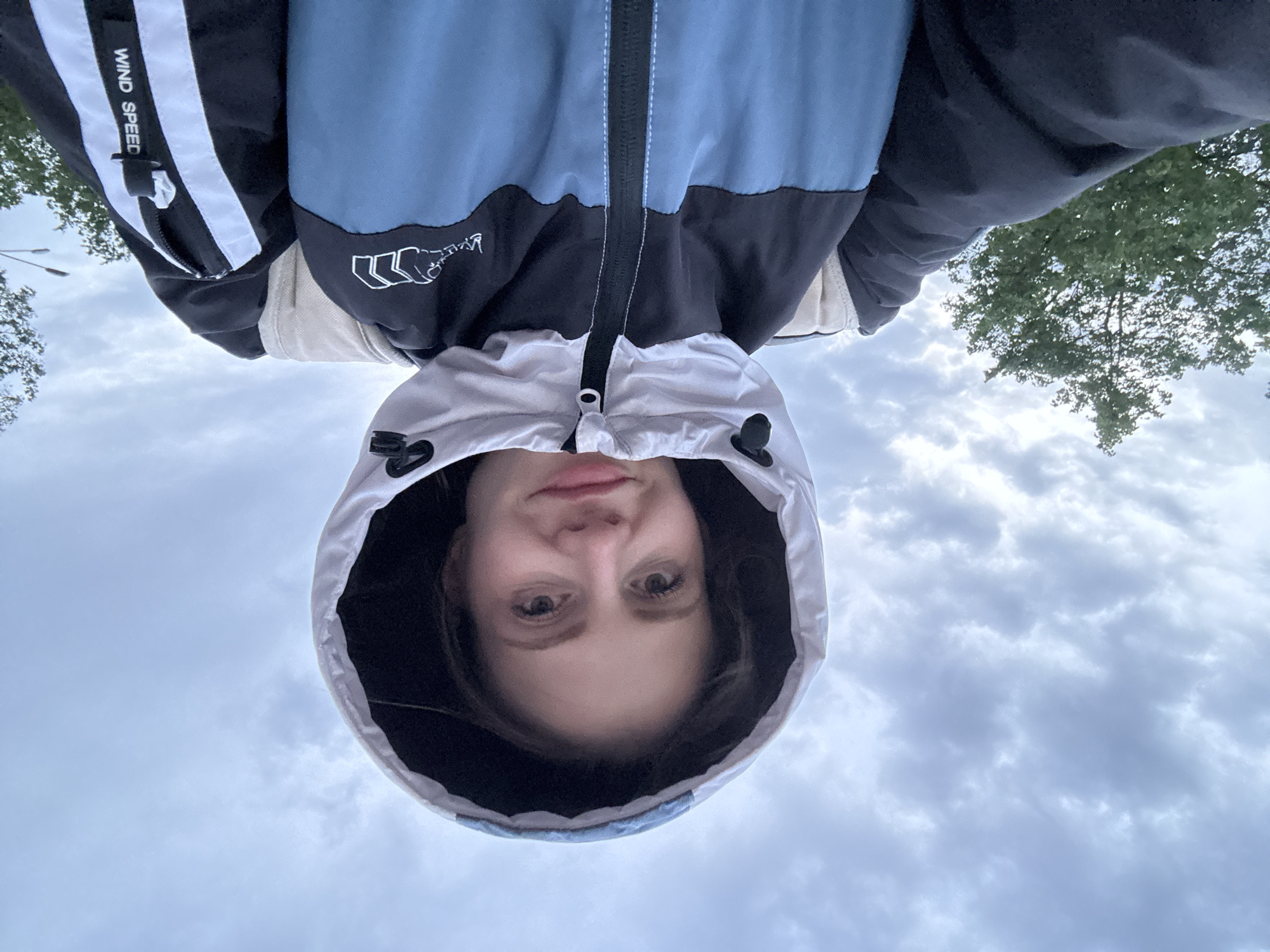 A person wearing a hooded jacket looks directly at the camera. The hood is pulled up, framing their face. The jacket is light blue and black with white accents. The background shows a cloudy sky and the tops of trees. Text on the jacket reads "WIND SPEED". Based on the style of architecture and vegetation, the location may be Saint Petersburg, Russia.