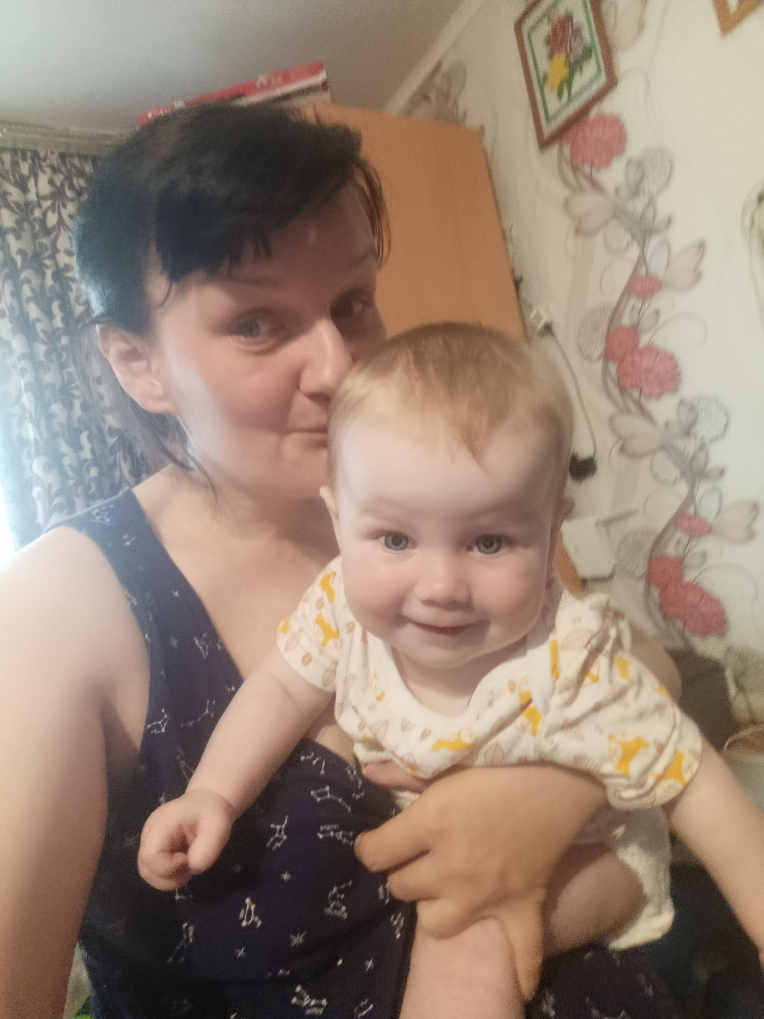 The image captures a warm and intimate moment between a woman and a baby in an indoor setting, likely a home in Фоки, Russia.
The primary subjects are a woman and an infant. The woman, positioned on the left side of the frame, has short dark hair and is wearing a dark blue sleeveless top adorned with white constellation-like patterns. Her face is turned towards the baby, and she appears to be kissing or nuzzling the baby's head, conveying an expression of deep affection. The baby, held securely in the woman's arms, has light blonde hair and bright blue eyes. The infant looks directly at the camera with a wide, joyful smile, revealing several small teeth. The baby is dressed in a white long-sleeve top with yellow animal patterns, and a white diaper is visible.
The activity taking place is a loving interaction between a mother (or caregiver) and a baby, captured in what appears to be a selfie. The emotions conveyed are happiness and tenderness.
The background details suggest a domestic setting. To the left, part of a light-colored wooden cabinet or cupboard is visible, with a red object (possibly a book or box) resting on top. Dark, patterned curtains hang behind the woman. The wall on the right is covered with patterned wallpaper featuring a light background adorned with delicate grey swirls and clusters of pink and red floral designs. A framed picture, displaying an embroidered or painted floral design with red and yellow flowers, hangs prominently on this wall. The lighting in the room is soft, suggesting indoor artificial light or an overcast day. No visible text is clearly discernible in the image.