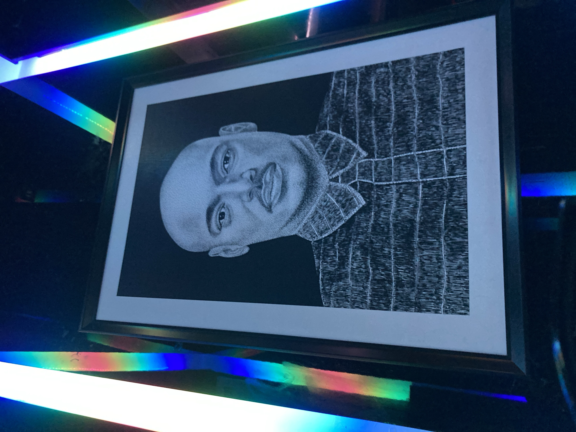 The image shows a framed black and white portrait of a bald Black man wearing a collared shirt. The portrait is on display, potentially in an art exhibition or a personal collection, and is illuminated by colorful, vibrant light sources that create a rainbow effect along the edges of the frame. The lighting suggests a modern or artistic setting. The specific location in Abuja, Nigeria, cannot be determined from the visual information provided in the image.