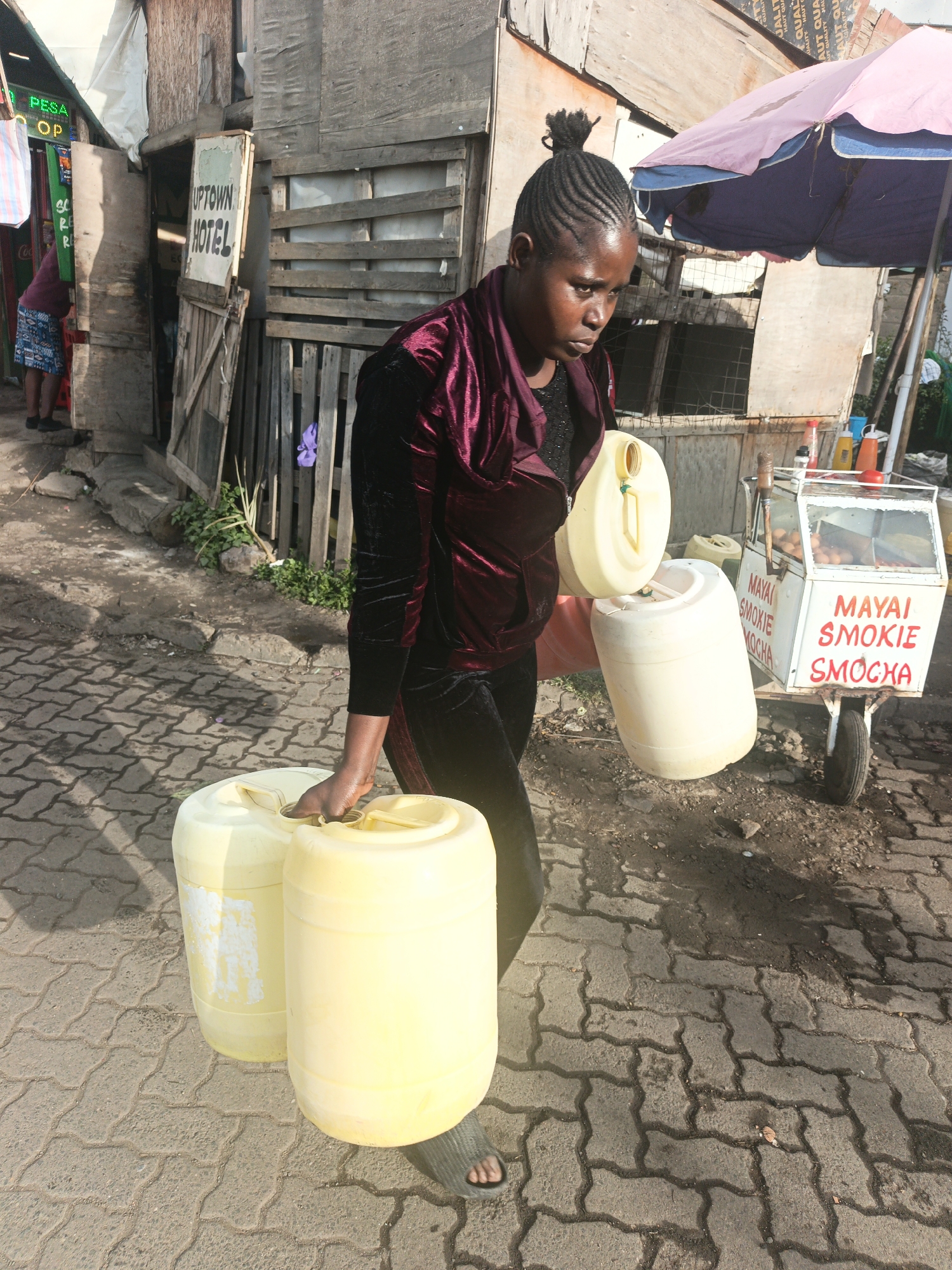 In Nairobi, Kenya, a woman carries three yellow plastic jugs along a cobblestone path. She wears a maroon velvet jacket and black pants, and sandals. Her hair is braided and pulled into a bun. Behind her is a weathered wooden structure with a sign reading "UPTOWN HOTEL." A small food cart labeled "MAYAI SMOKIE SMOCHA" stands nearby under a blue and pink umbrella. A person is visible in the background near a sign with "PESA OP" printed on it. The ground is paved with interlocking gray stones.