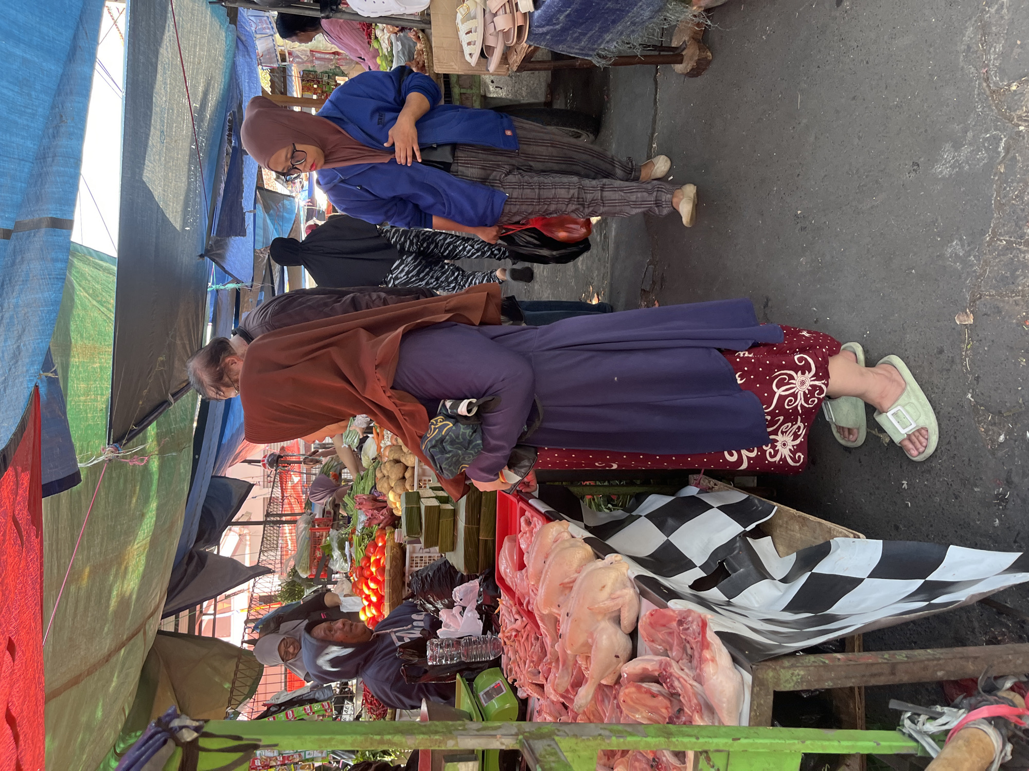 This image depicts an outdoor market scene in Bandung City, Indonesia. In the foreground, raw chicken is displayed on a table covered with a black and white checkered cloth. To the right, a woman wearing a blue jacket, plaid pants, and a brown hijab is holding a red plastic bag. Behind her, a young woman with glasses and a brown hijab is looking towards the left. The market stalls are covered with colorful tarps in red, green, and blue. In the background, other market-goers and produce, including tomatoes and potatoes, are visible. The ground is paved with asphalt.