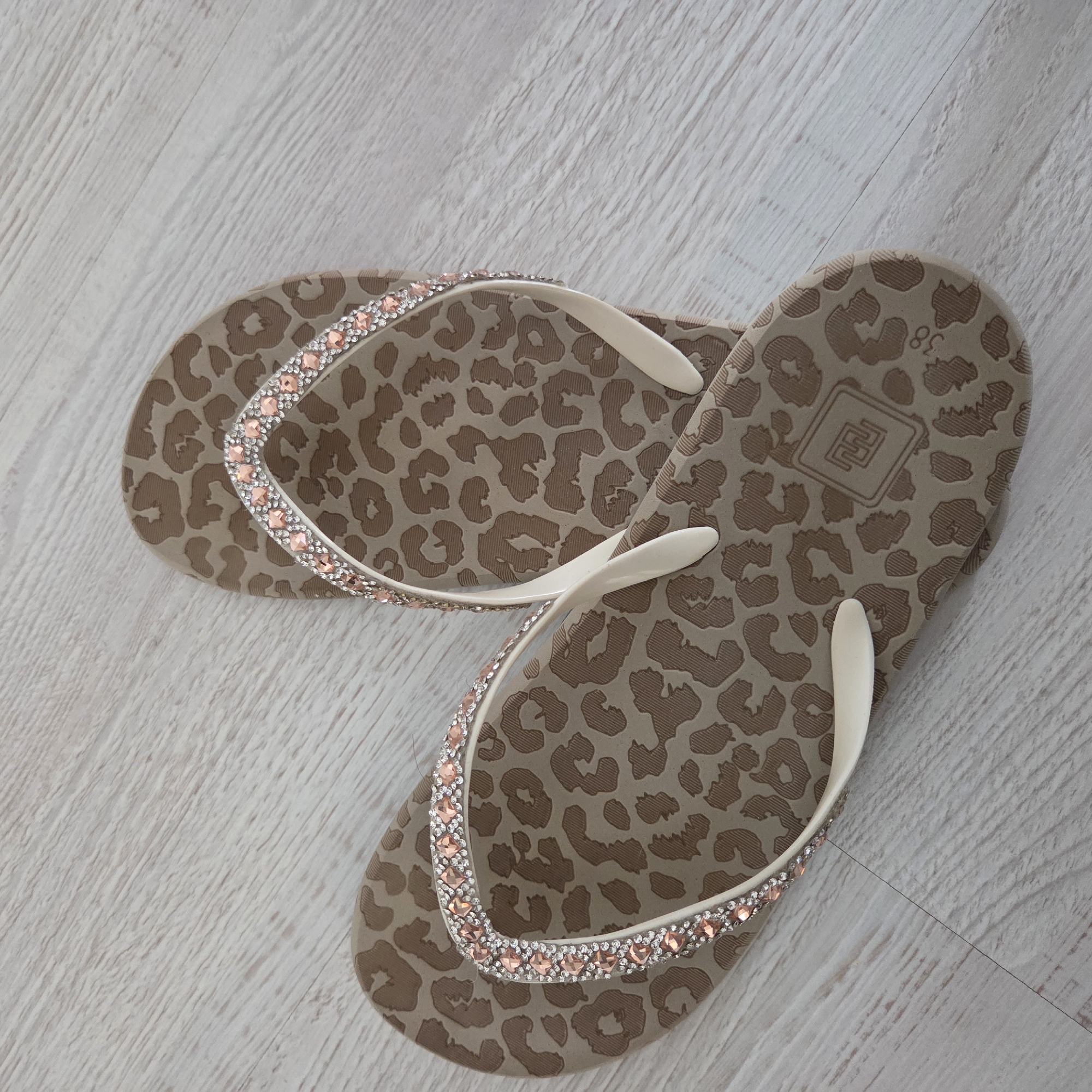 A pair of women's flip-flops is resting on a light-colored wooden or laminate floor, viewed from directly above. The flip-flops are primarily beige/tan with a debossed or embossed leopard print pattern covering the entire surface of their soles. The straps are made of a cream or off-white material.
Both straps are embellished with two rows of sparkling, square-cut rhinestones. The inner row consists of smaller, clear or silver-toned rhinestones, while the outer row features slightly larger, pink or peach-colored rhinestones. The right flip-flop is positioned above and slightly to the left of the left flip-flop, creating a somewhat diagonal arrangement.
On the heel area of the right flip-flop's sole, the number "38" is visible, likely indicating the shoe size. Below the "38" and towards the center of the heel, there is a square logo with a stylized geometric pattern inside, which appears to be a brand mark. The lighting is even, suggesting an indoor setting with either natural ambient light or soft artificial lighting. No other objects or subjects are visible in the frame.
