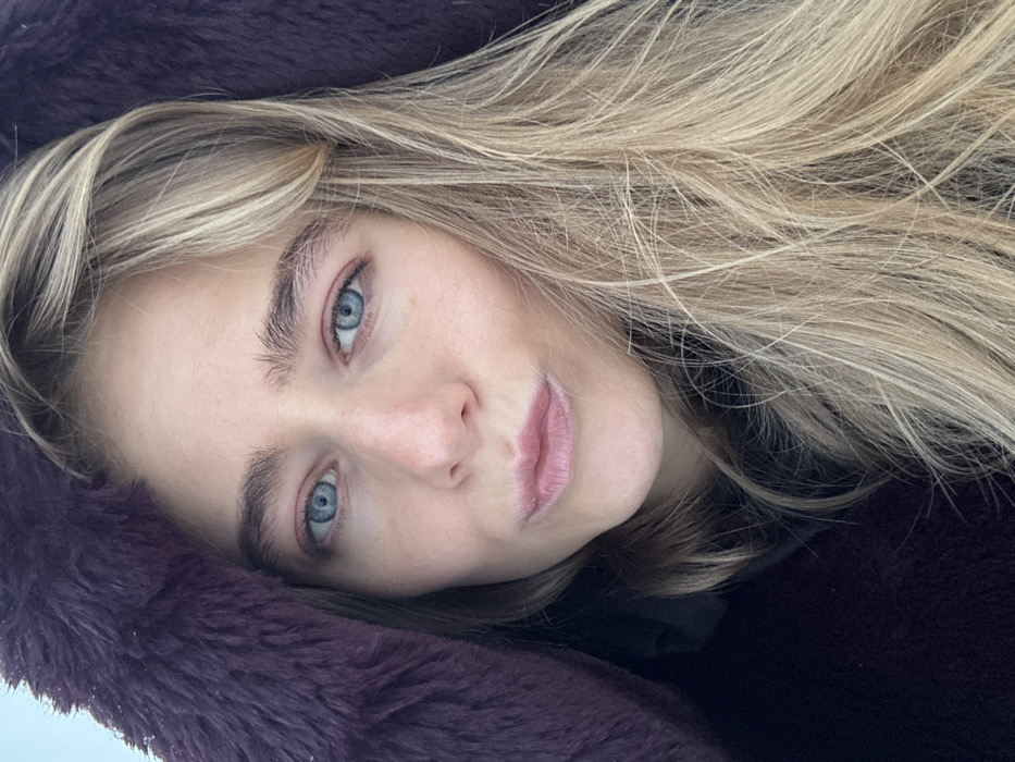 A close-up, head-and-shoulders portrait depicts a young adult female, located in the city of Unknown, Unknown. The subject has fair skin, blue irises, and long, wavy blonde hair with lighter highlights, extending down the right side of the frame. Her eyebrows are light brown and appear untweezed. Her lips are slightly pursed in a gentle pout. She is wearing a dark garment, possibly a hoodie, underneath a dark purple or burgundy faux fur-lined hooded coat. The faux fur trim of the hood is visible along the left side of her face. The background is out of focus, appearing as a bright, indistinct expanse, consistent with an outdoor, possibly overcast or snowy environment. The scene is illuminated by soft, natural light.