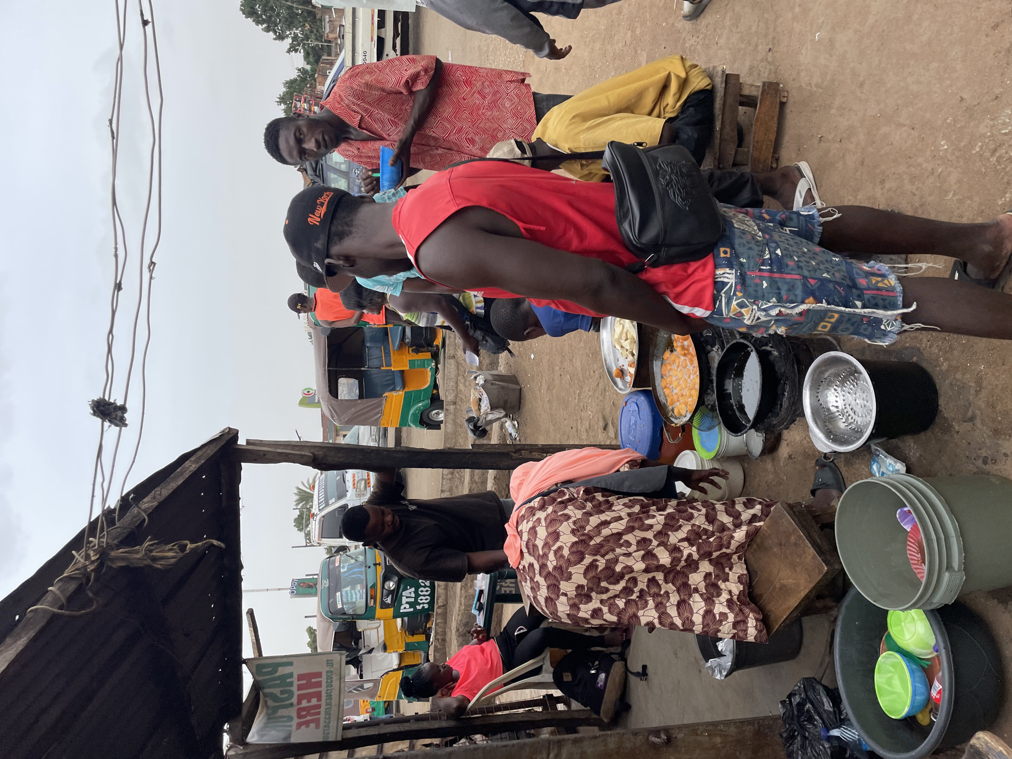 The image shows a busy street scene in Jos, Nigeria, with several people gathered around a makeshift food stall. The stall is under a rough wooden structure with a metal roof, and a sign is visible with the word "HERE." Multiple auto rickshaws (tuk-tuks) are parked nearby.
Several individuals are visible: one man in a red shirt and patterned shorts stands near the stall, another man in a red patterned shirt sits on a wooden structure, and other figures are partially obscured in the background. A woman sits in a chair at the edge of the image, wearing a pink shirt.
The food stall displays an array of metal pots, buckets, and trays containing food items, likely for sale. Some of the items appear to be fried snacks.