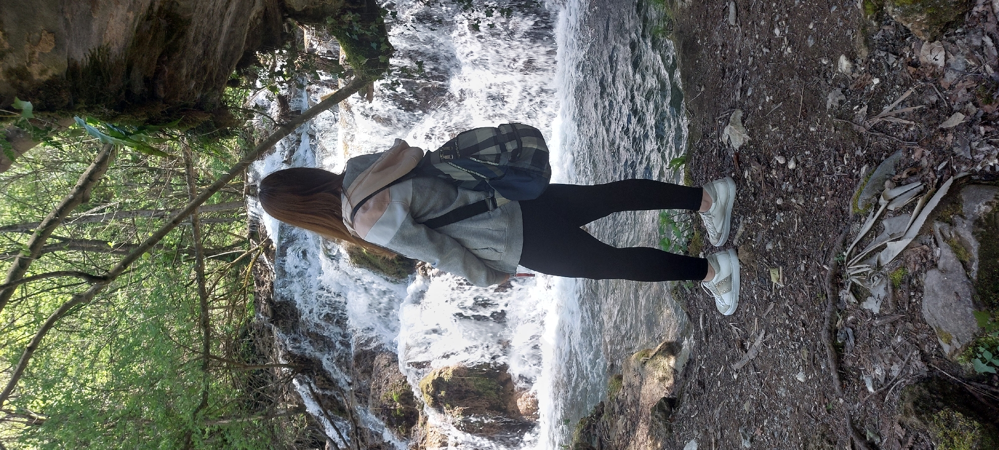 A young woman stands on a dirt path next to a waterfall at Бачковски манастир, Bulgaria. She is wearing black leggings, white sneakers, a grey hoodie with a beige section on the back, and a dark-colored backpack. The waterfall is to her left, cascading over rocks and forming a stream. Lush green trees and vegetation surround the area, with a rock formation visible in the upper left corner. The dirt path is covered with leaves, small rocks, and roots.