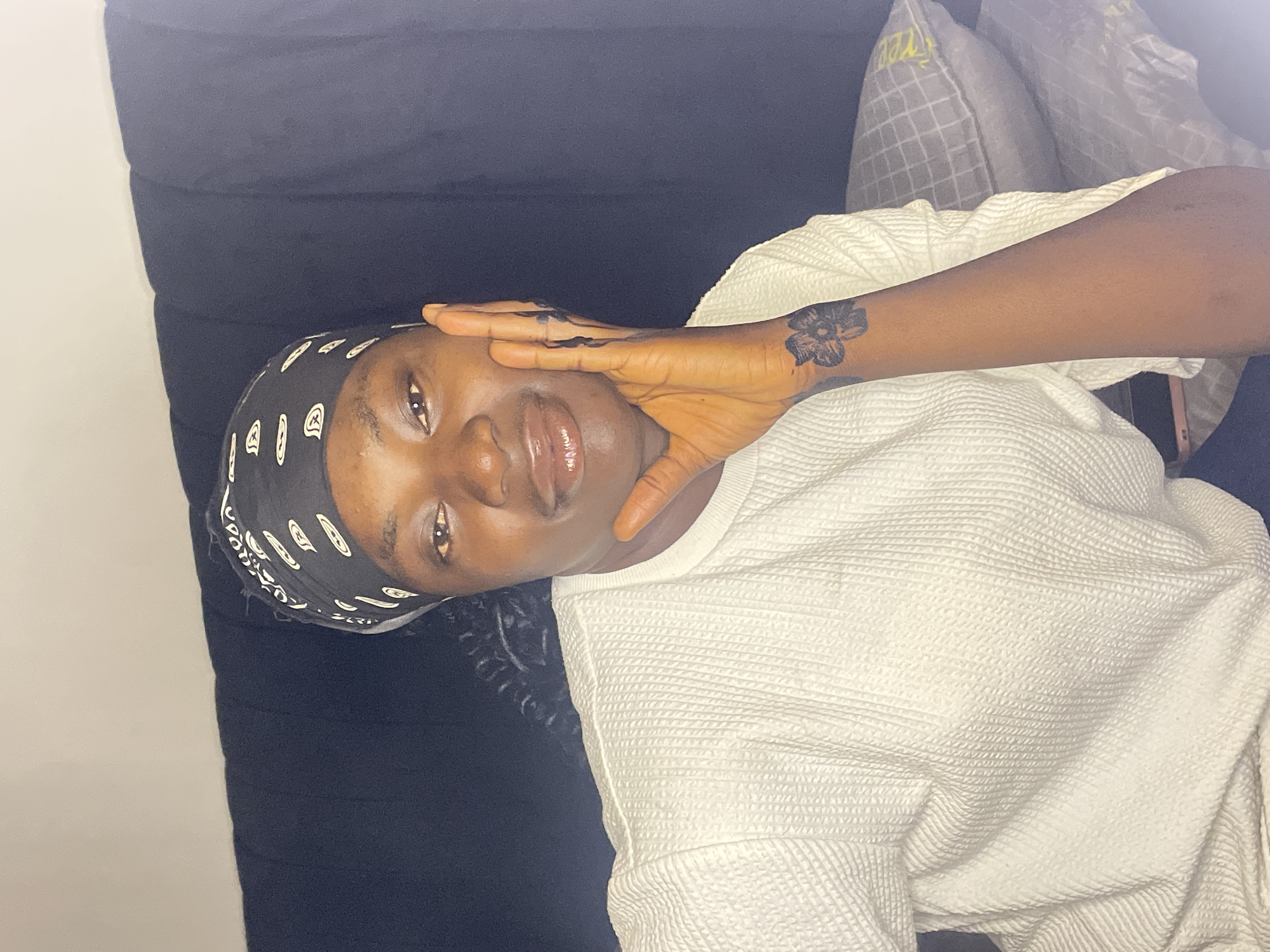 A young individual, likely female, from Jos, Nigeria, is captured in a close-up, rotated portrait, appearing to lie down on a dark surface, possibly a bed or couch. Her head is turned towards the camera, with her right hand gently resting on the side of her face. She has a serene expression, with glossy lips and open eyes looking directly at the viewer.
She is wearing a black headscarf or bandana adorned with white, abstract patterns. Her attire includes a white, long-sleeved top with a distinct waffle-knit or textured pattern. A notable detail is a black flower-shaped tattoo on the back of her right hand or wrist.
In the background, behind her head, are two pillows. One has a light grey grid pattern with faint yellow text partially visible, which appears to spell "YEE". The setting seems to be indoors, with soft, indirect lighting. The overall mood conveyed is calm and relaxed.
