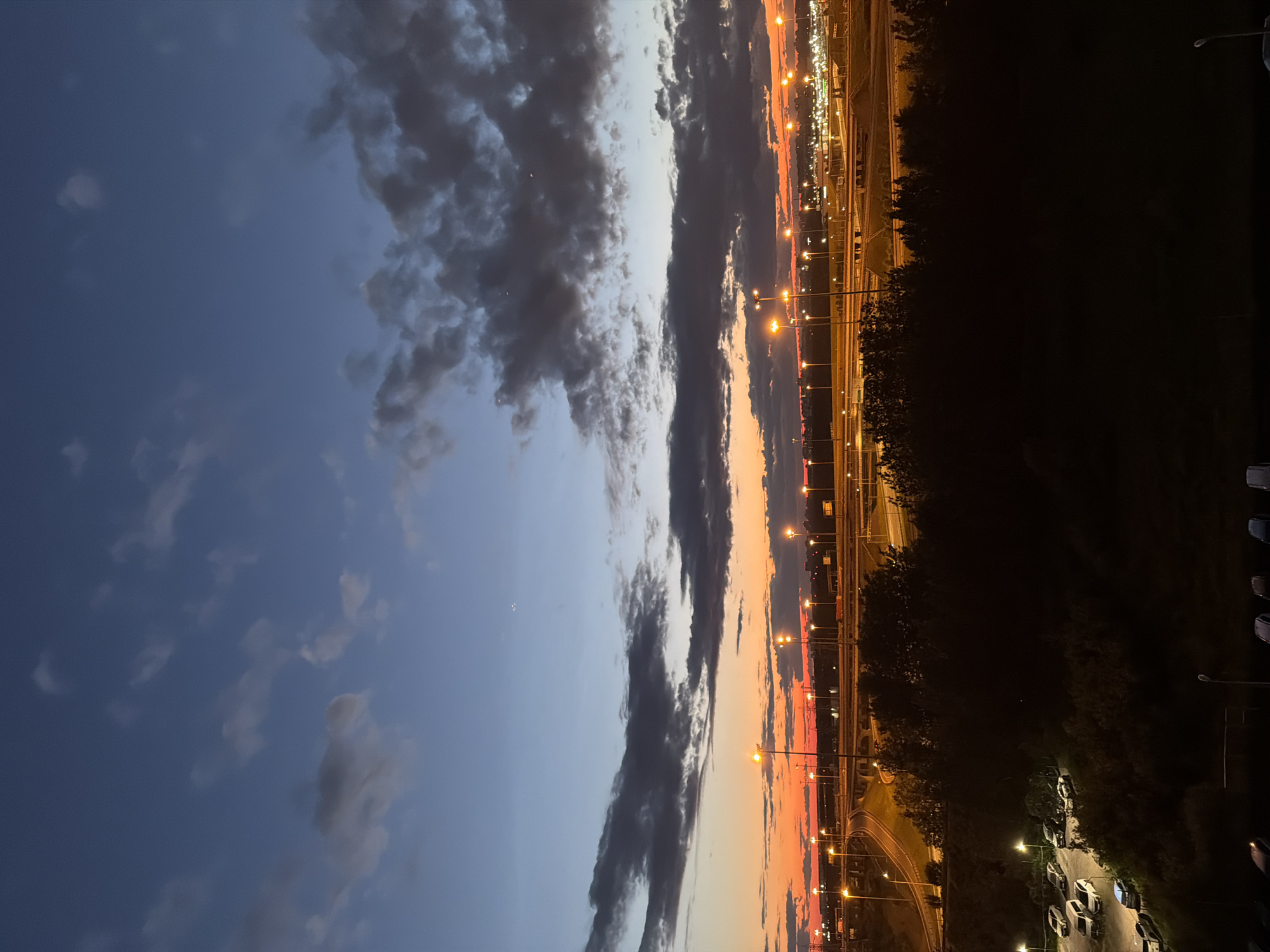 A wide-angle, high-angle shot captures a sunset over a city with an elevated highway system. The sky transitions from a deep blue at the top to a vibrant orange and pink hue near the horizon, with scattered clouds. Numerous streetlights illuminate the highway and surrounding areas, creating a network of glowing dots against the twilight sky. In the foreground, a dark silhouette of trees and a parking lot filled with cars is visible. The overall scene depicts an urban landscape transitioning from day to night.