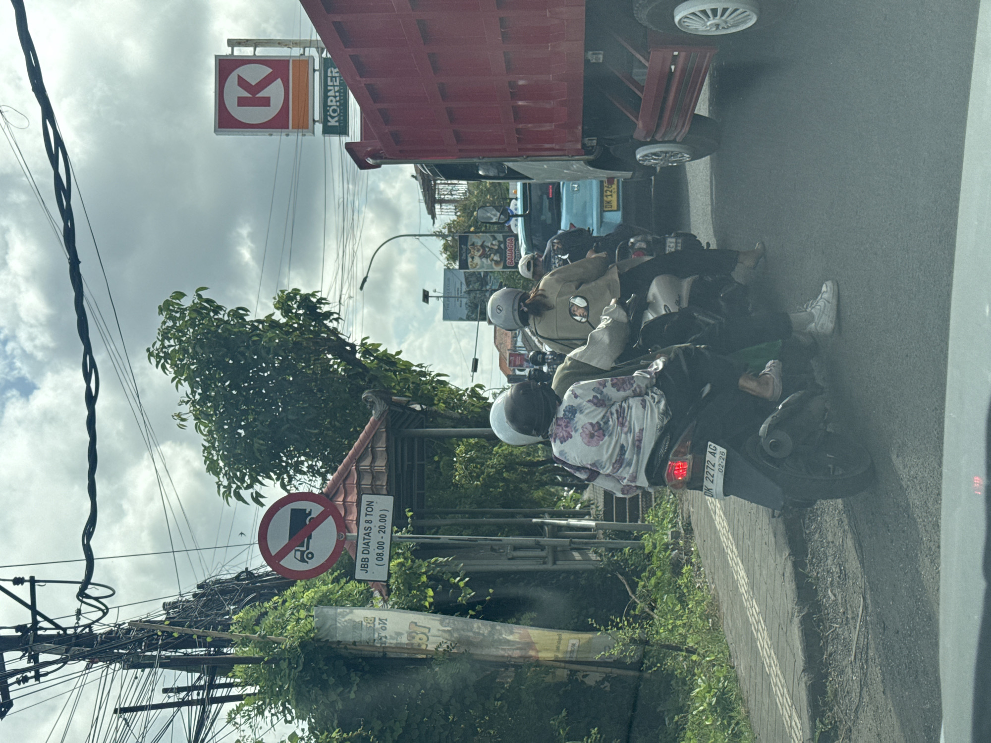 The image depicts a busy street scene in Kuta Selatan, Indonesia. Several scooters are stopped in traffic behind a red truck with a "Korner" convenience store sign visible above. A blue car with the license plate "DK 126" is also visible. There are utility poles with tangled wires on the left side of the frame, and a sign indicating "JBB DIATAS 8 TON (08.00-20.00)" which translates to "Weight above 8 tons prohibited from 8:00 AM to 8:00 PM." One scooter has the license plate "DK 2272 AG 02-26." The weather is overcast. There are various commercial signs and banners present. The overall impression is one of urban congestion.