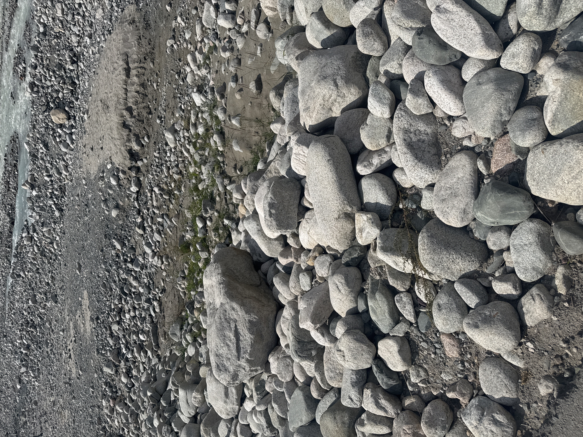 An overhead, medium shot captures a vast expanse of a rocky riverbed in an outdoor, natural environment. The riverbed is densely packed with a multitude of stones, varying in size, shape, and color from light gray to darker, mottled tones. Some larger, boulder-like rocks are scattered throughout, while smaller pebbles and gravel fill the spaces between. In the upper right quadrant of the image, a body of flowing water, likely a river, is visible, its surface agitated with ripples. A patch of sandy earth with sparse, dry grass emerges from the stones in the mid-ground. The overall scene suggests a wilderness area, possibly in a mountainous region, given the rocky terrain and flowing water. The lighting is natural, indicating daylight, casting subtle shadows among the rocks. There are no people or animals present in the frame. The image appears to be taken from a slightly elevated position, looking down at the riverbed. The scene evokes a sense of raw, natural landscape, untouched by human intervention, typical of remote natural areas.