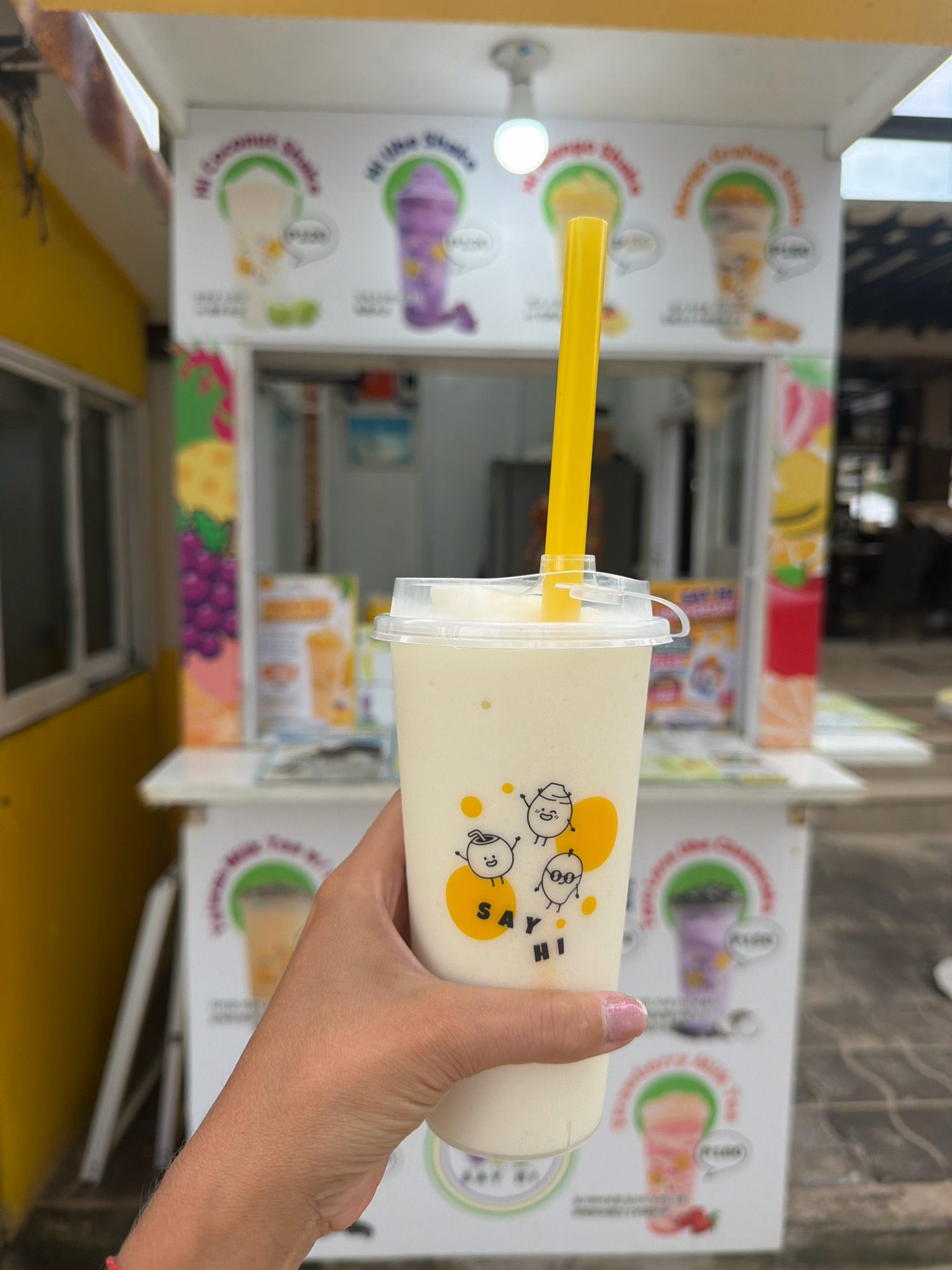 A person's hand, with pink nail polish on the ring finger, holds a clear plastic cup filled with a pale yellow smoothie. The cup has a white lid and a yellow straw protruding from it. The cup is decorated with cartoon drawings of round, smiling fruits or vegetables. In the background, there is a brightly colored food stall with various drink advertisements displayed on its front. The stall appears to be outdoors, with a yellow building to the left. One advertisement shows a purple drink, another a yellow drink, and a third a pink drink with strawberries. The overall scene suggests a casual, outdoor dining or beverage stand in Malay, Philippines.