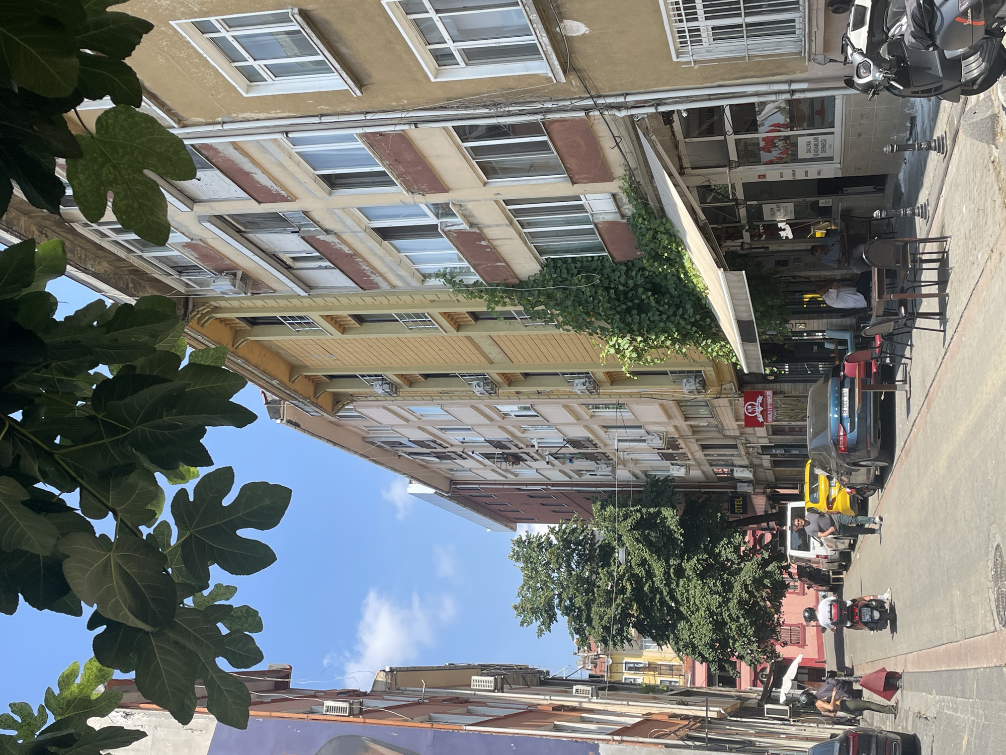 The image shows a street scene in Kadıköy, Turkey, on a sunny day. The street is lined with multi-story buildings on both sides. The buildings have a mix of architectural styles and colors, with some appearing older and others more modern. In the foreground, there are trees with large leaves partially obscuring the view of the sky.
On the left side of the street, there is a building with a purple and orange facade. Further down the street, a large green tree is visible between the buildings. There are several parked cars, including a yellow taxi, a white van, and a blue sedan. A person is riding a scooter, and another person is walking down the street. There are also several tables and chairs set up outside a cafe on the right side of the street, with a few people sitting at them.
The street is paved with cobblestones, and there are streetlights and power lines visible. The sky is blue with a few scattered clouds. The overall impression is of a bustling urban environment in a Mediterranean or similar climate.