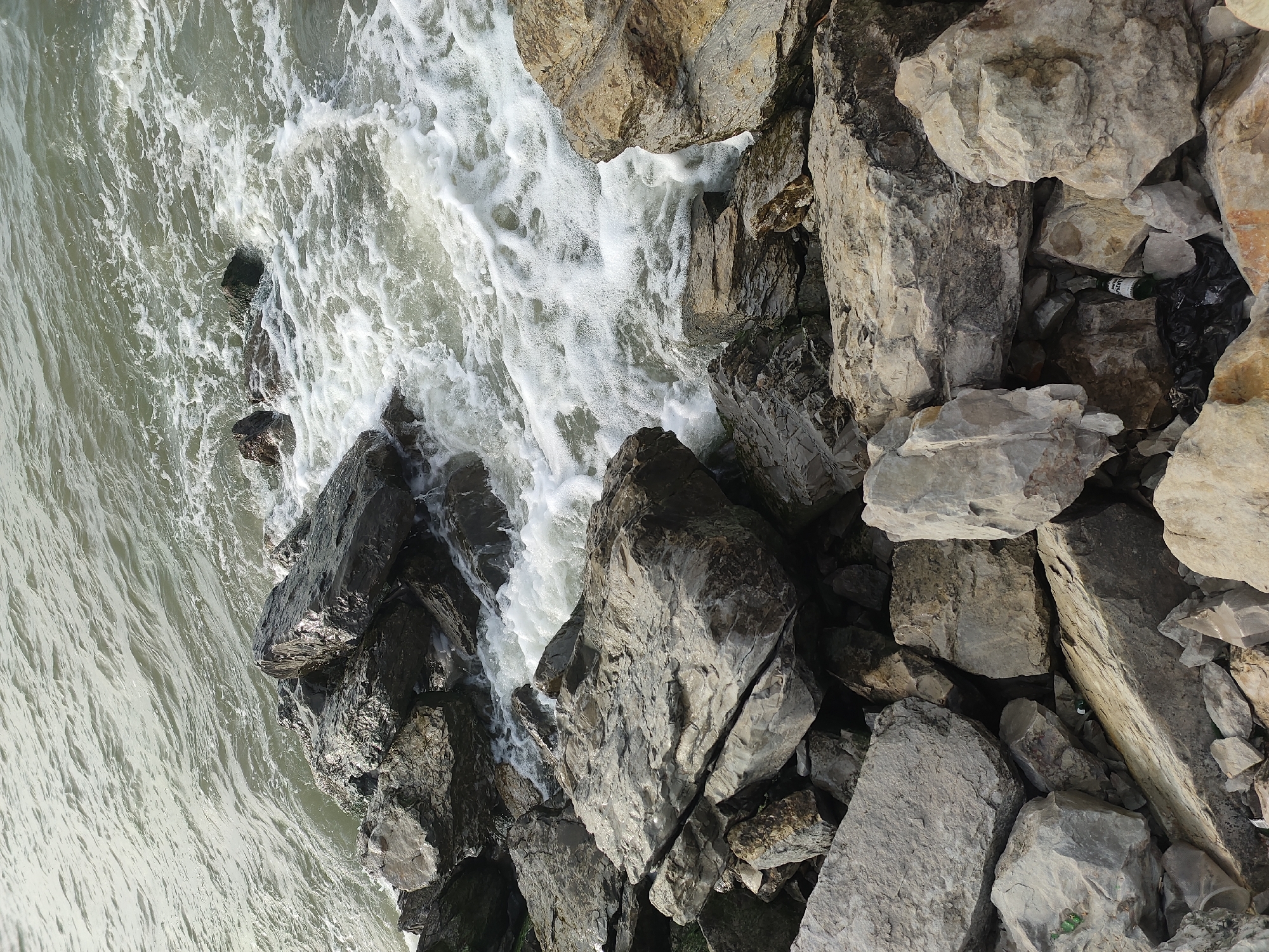 The image shows a rocky coastline with waves crashing against the shore. The water is choppy and foamy, and the rocks are dark and wet. There is a small amount of trash visible amongst the rocks, including a plastic bag and a green glass bottle. The scene appears to be natural and untouched, with the exception of the litter. It is difficult to ascertain the exact location in Makhachkala, Russia, based solely on the image.