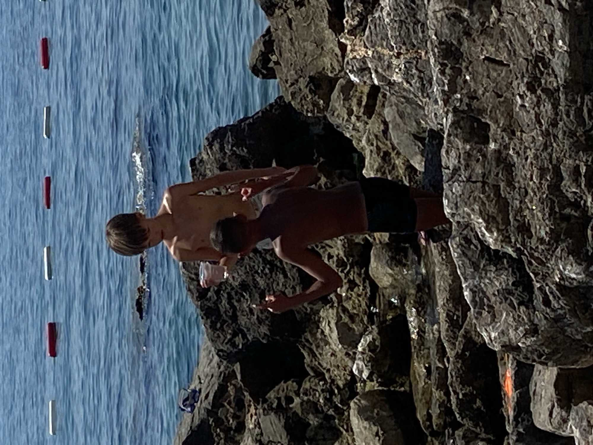 The image depicts two young males on a rocky coastline with the blue sea in the background. One person, wearing dark swim trunks, is positioned on the rocks, looking down and holding a clear cup with a reddish liquid. The other person, with lighter hair, is also on the rocks, facing towards the sea and holding a phone. They appear to be at a beach with buoys marking a swimming area. The lighting suggests it is daytime, possibly late afternoon, with the sun casting shadows on the rocks. The scene evokes a relaxed, summery atmosphere. The location is identified as Prijevor, Montenegro. There is no visible text in the image.