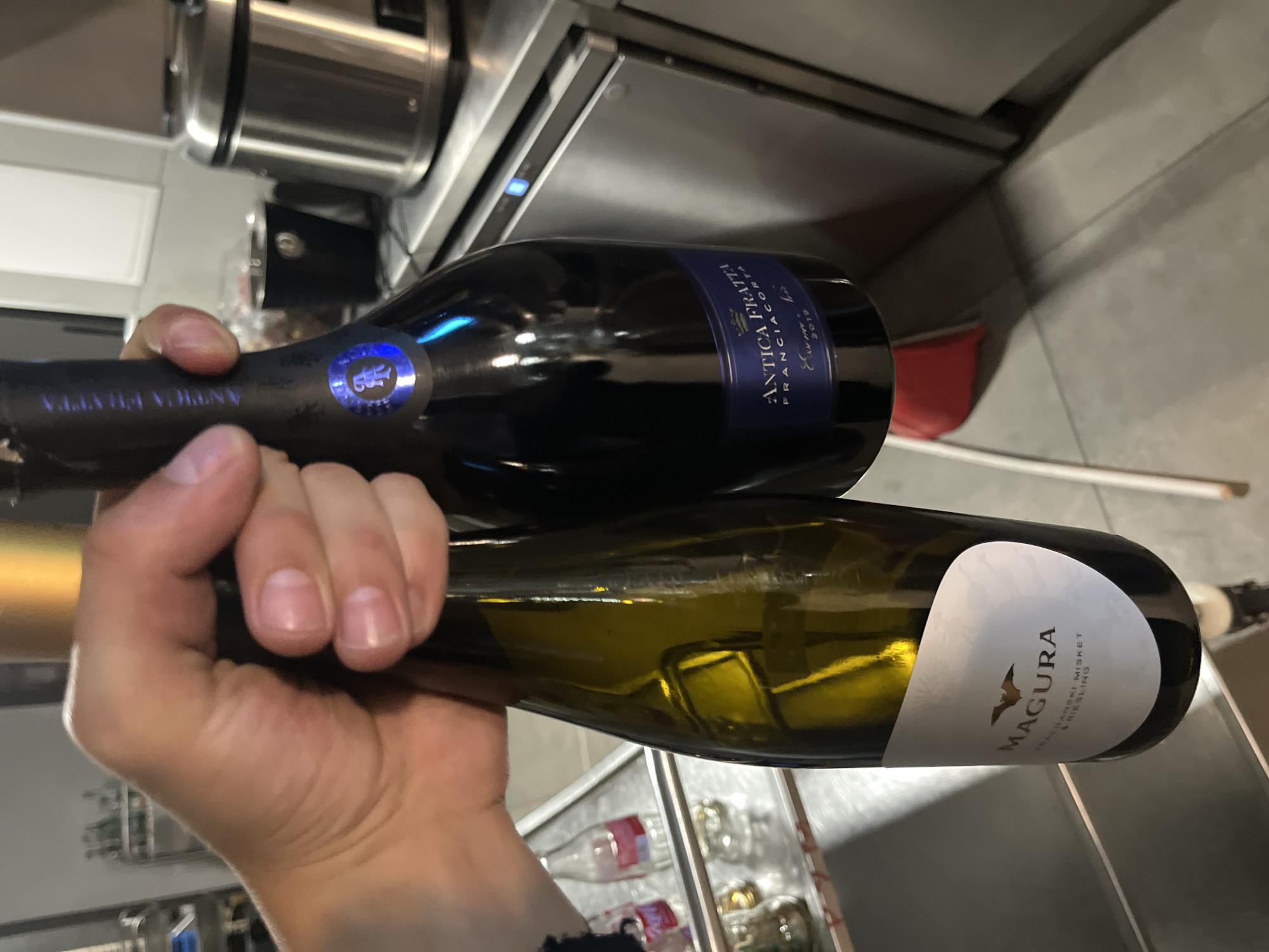 A person is holding two bottles of wine. The top bottle is dark and labeled "ANTICA FRATTA Franciacorta," while the bottom bottle is lighter in color with a white label that reads "MAGURA." The bottles are held against a backdrop of a stainless steel kitchen area. There is no indication of the city of Sofia or Bulgaria present in the image.