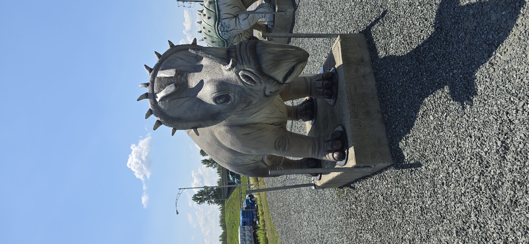 This is a photograph of a dinosaur statue, likely part of an outdoor exhibit. The statue is of a triceratops, depicted with its head lowered, appearing to be walking. It is made of a grey, textured material resembling concrete or fiberglass, and is mounted on a concrete base. The statue has a large frill around its head adorned with spikes, and three horns. Its eyes are visible, and its feet are detailed with claws. Metal poles and chains are attached to the statue, possibly for support or to secure it.
In the background, another dinosaur statue, a stegosaurus with a green back and plates, is partially visible. The setting is outdoors, with a clear blue sky and scattered white clouds. The ground is covered in small grey gravel. To the right of the triceratops, a large shadow is cast, indicating it is daytime, likely mid-morning or afternoon due to the angle of the sun. Further in the background, there are trees, a road with cars and a blue truck, and a lamppost. The location is identified as Saint-Léonard-d'Aston, Canada. The overall impression is a sunny day at an outdoor attraction. There is no visible text in the image.