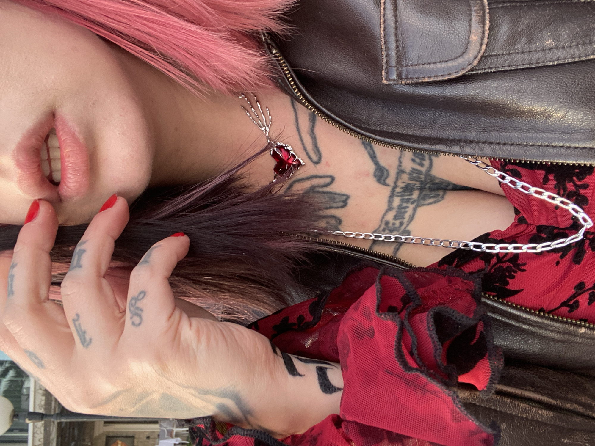 A close-up image captures the upper torso of an individual in Rafailovići, Montenegro, from the chest to just below the eyes. The subject's right hand, bearing multiple dark finger tattoos and a larger wrist tattoo, is positioned with red-painted fingertips near the mouth. The lips are slightly parted, revealing upper and lower teeth. Hair is bi-colored, dark on the viewer's right and bright pink on the viewer's left. The individual wears a partially unzipped dark brown leather jacket over a red garment with black floral patterns and ruffled details. Two silver-colored necklaces are visible: one features a skeleton hand pendant holding a red heart, and a thicker chain hangs lower. Multiple dark tattoos, including script, are visible on the chest. The out-of-focus background depicts an outdoor setting with a white building, a dark lamp post, and faint outlines of tables and chairs, illuminated by soft daylight.
