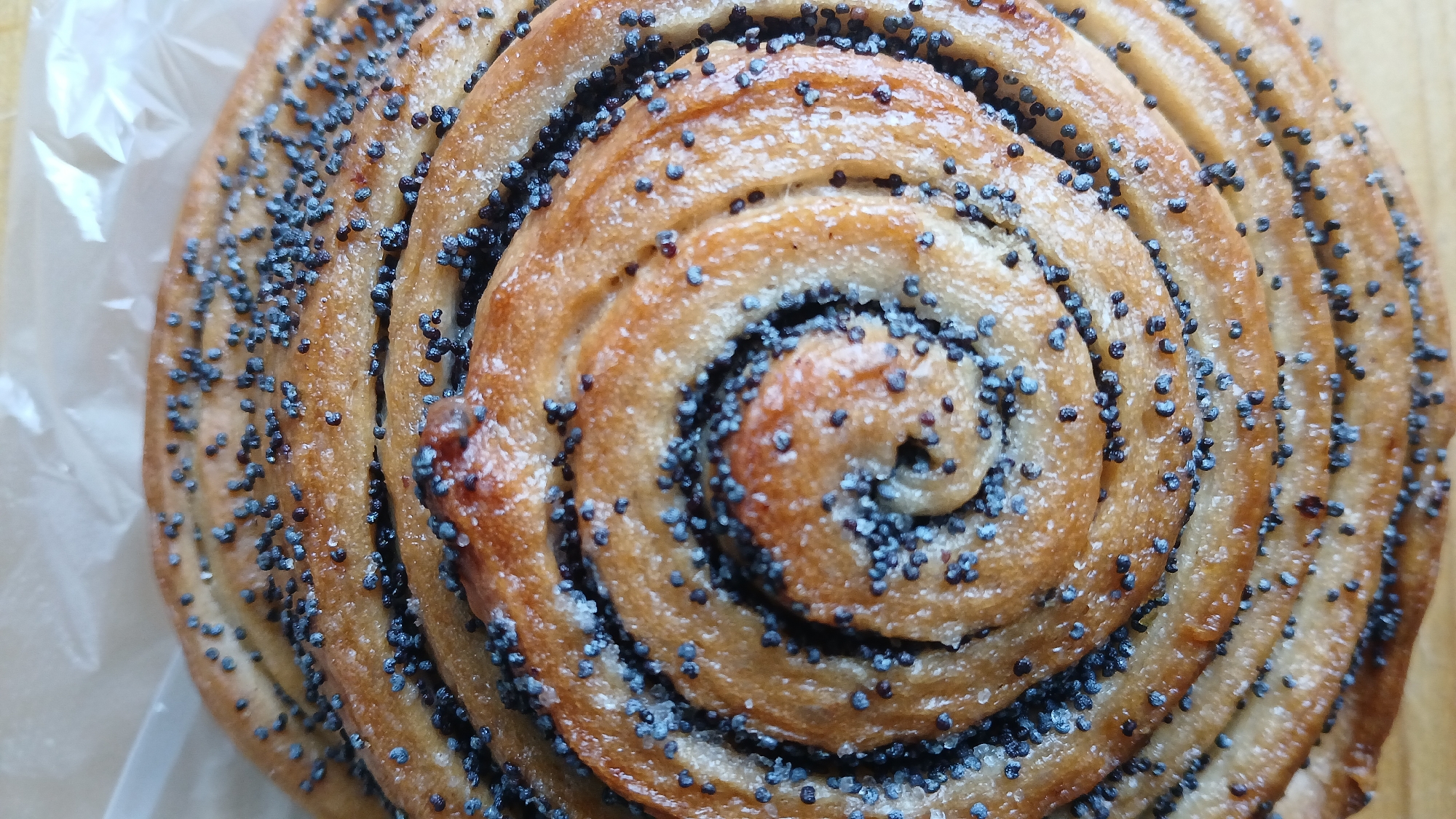 A close-up, overhead shot of a swirled pastry dusted with poppy seeds rests on a piece of white paper. The pastry exhibits a golden-brown hue with visible layers, and the poppy seeds are scattered across its surface, with a denser concentration on the outer layers. The background is out of focus, showing a hint of a wooden surface. This image does not depict any specific location in the city of Rubtsovsk, Russia.