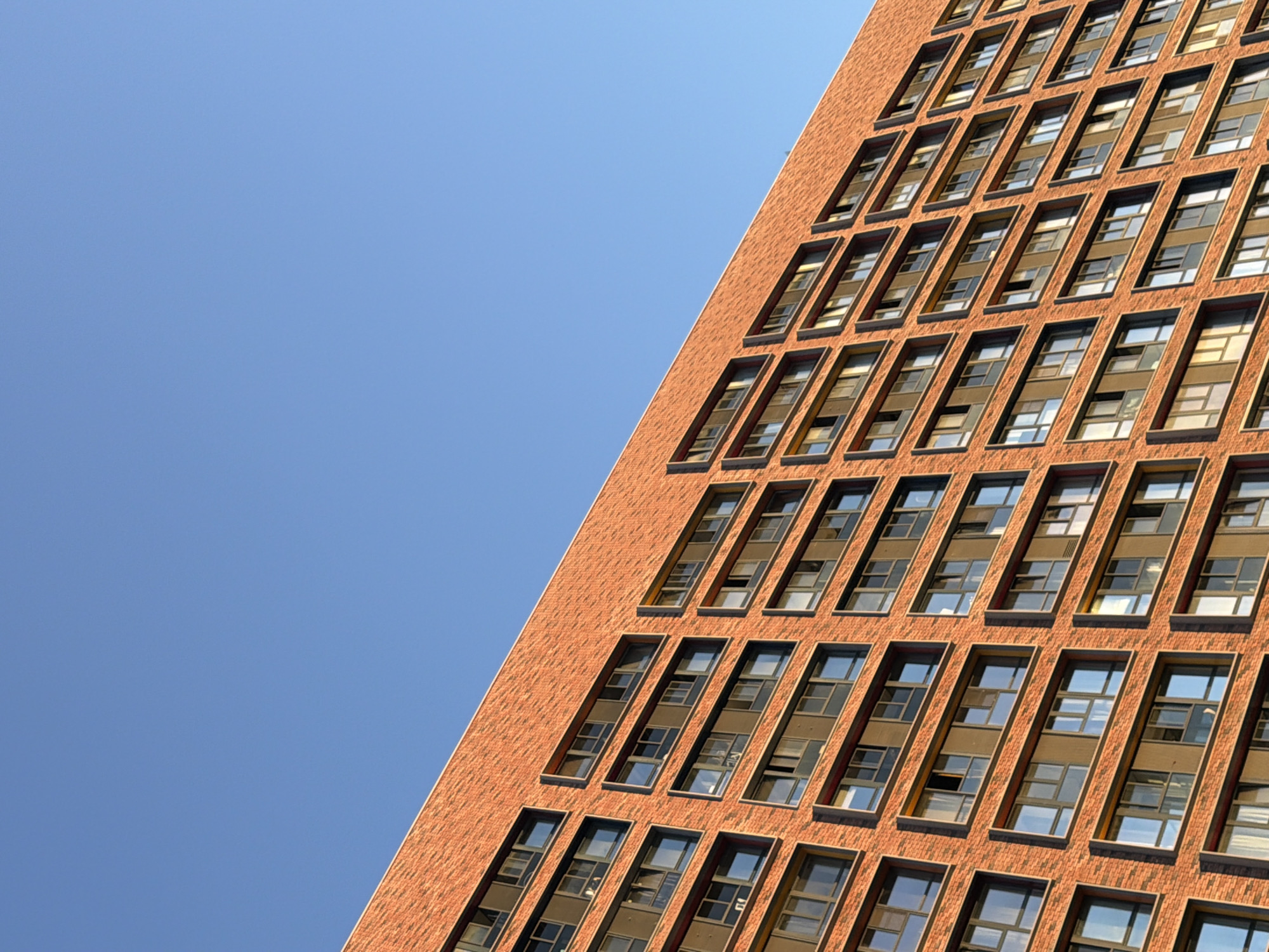 The image is a close-up, angled shot of the exterior of a modern, multi-story brick building. The building features a repeating pattern of dark-framed windows. The brickwork is a warm orange-red color, and the texture is visible, appearing as small, uniform bricks. The windows are recessed and appear to be set into darker frames. The sky above is a clear, bright blue, occupying the upper portion of the frame. The lighting suggests it is daytime, likely with the sun casting a warm glow on the building. The perspective is from a low angle, looking up at the building. There are no people or other distinguishing features in the frame.