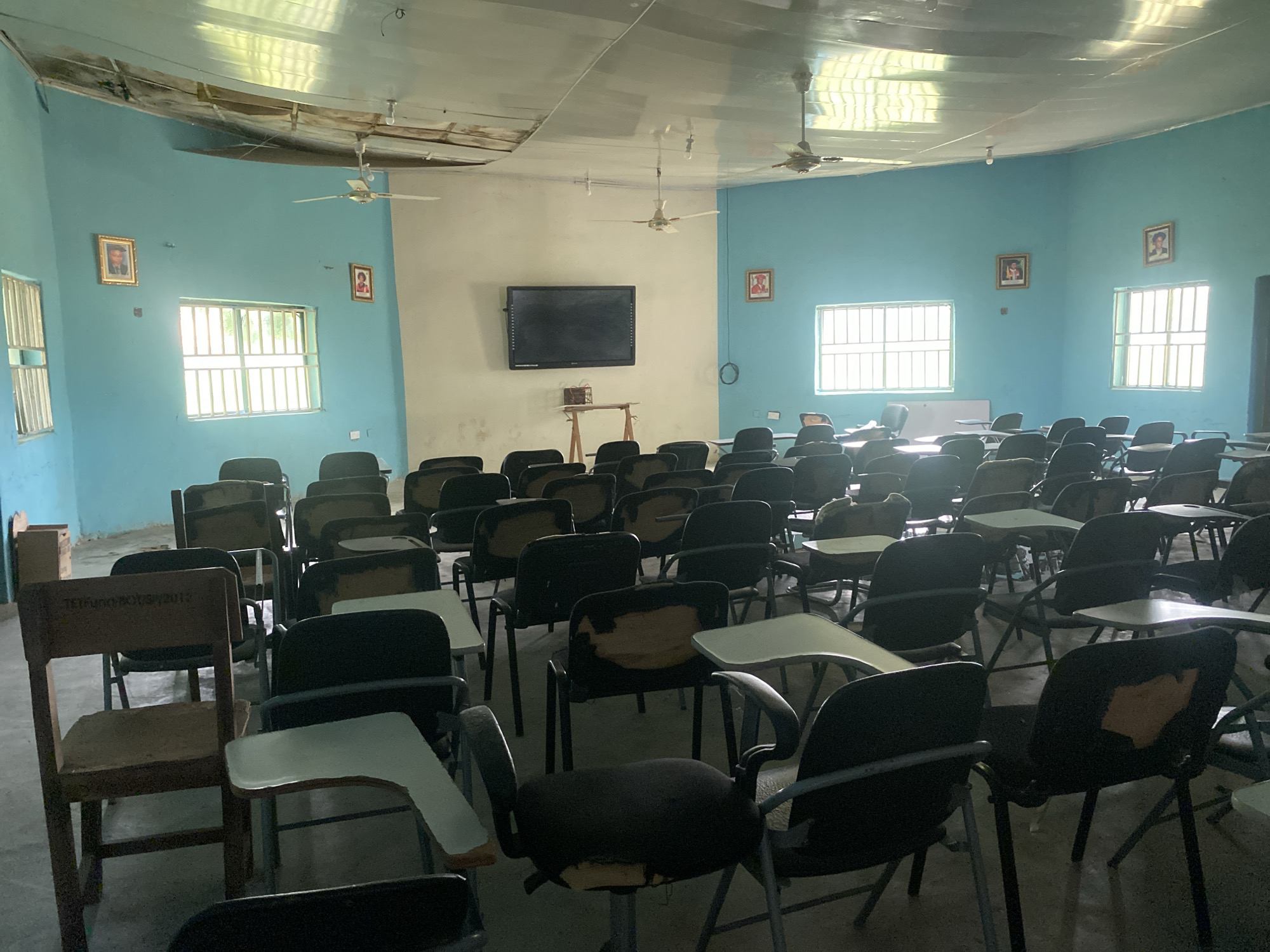 The image shows an empty classroom in Jalingo, Nigeria. The room has pale blue walls and a reflective ceiling that shows signs of damage. Several ceiling fans are visible, some of which appear to be older models. The classroom is furnished with rows of desks and chairs, many of which are worn or damaged. Some of the chairs have ripped upholstery, and some desk surfaces are chipped or stained. Two framed pictures hang on the walls, and a large flat-screen television is mounted on the wall at the front of the room, above a small table. Sunlight streams in through barred windows, casting shadows across the room. The overall impression is one of disuse or neglect.