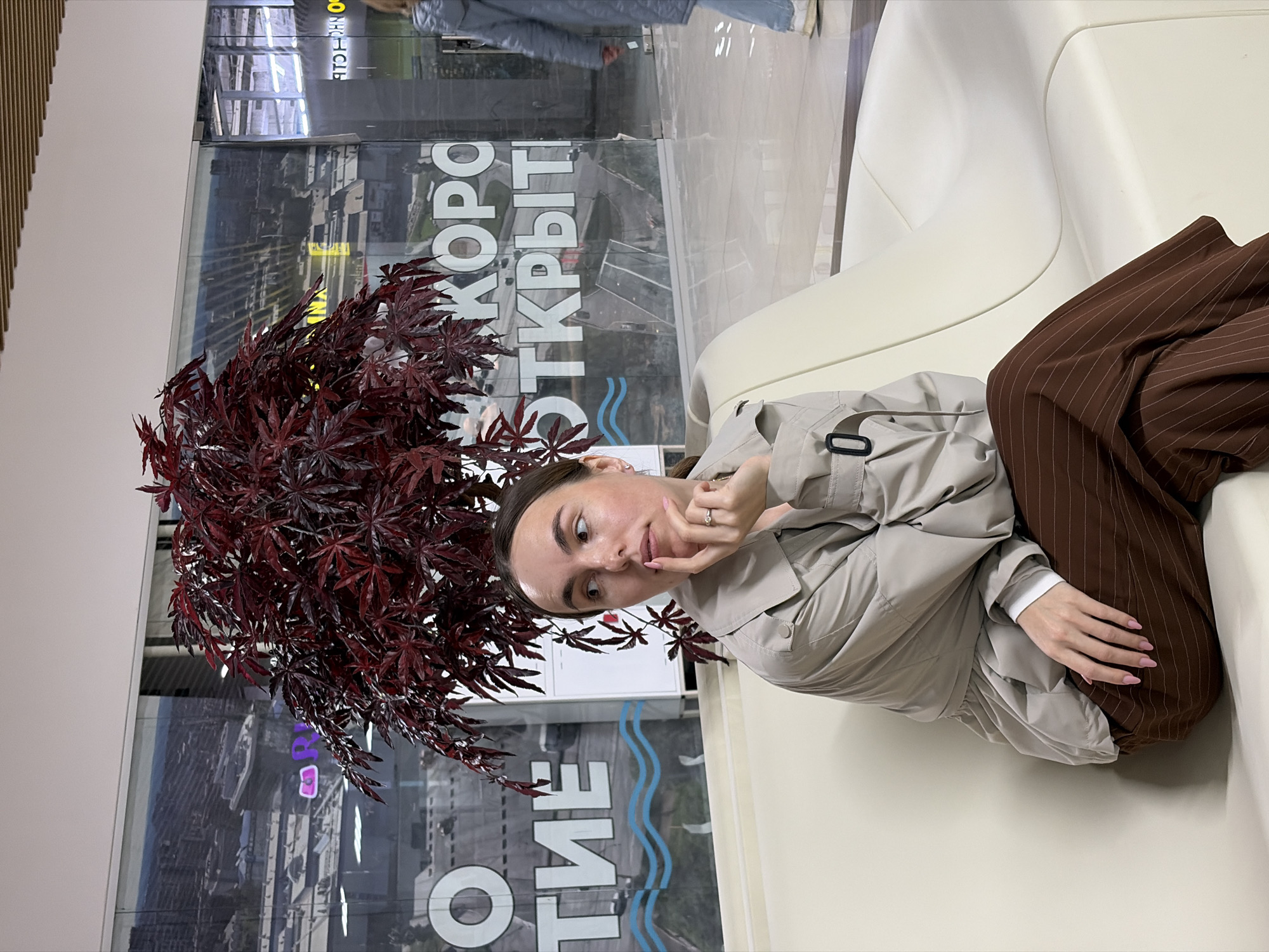 The image, taken in Izhevsk, Russia, features a young woman sitting on a cream-colored upholstered bench or sofa in an indoor public space, likely a shopping mall or commercial area. The photo is rotated 90 degrees clockwise.
The woman is wearing a light beige or greyish-tan trench coat or jacket and dark brown trousers with thin white pinstripes. She has dark hair pulled back and is looking towards the camera with a slightly thoughtful or playful expression, her left hand lightly touching her chin/mouth, displaying a ring on her ring finger and pink nail polish.
To her left, a prominent plant with striking deep red, palmate leaves, resembling a Japanese maple, stands as a decorative element. The dense foliage of the plant creates a vibrant contrast.
Behind the woman and the plant is a large glass partition or window. Reflected on this glass, mirrored text is visible, which reads "СКОРО ОТКРЫТИЕ" (SKORO OTKRYTIYE), translating to "COMING SOON" or "OPENING SOON". Below this text, a distinctive blue wavy logo can be seen. The reflections in the glass also show glimpses of the surrounding indoor environment and possibly blurred exterior views. The setting appears modern and well-lit, suggesting an active commercial space.
The woman seems to be posing for a photograph. The scene depicts an ordinary moment in a public setting.
