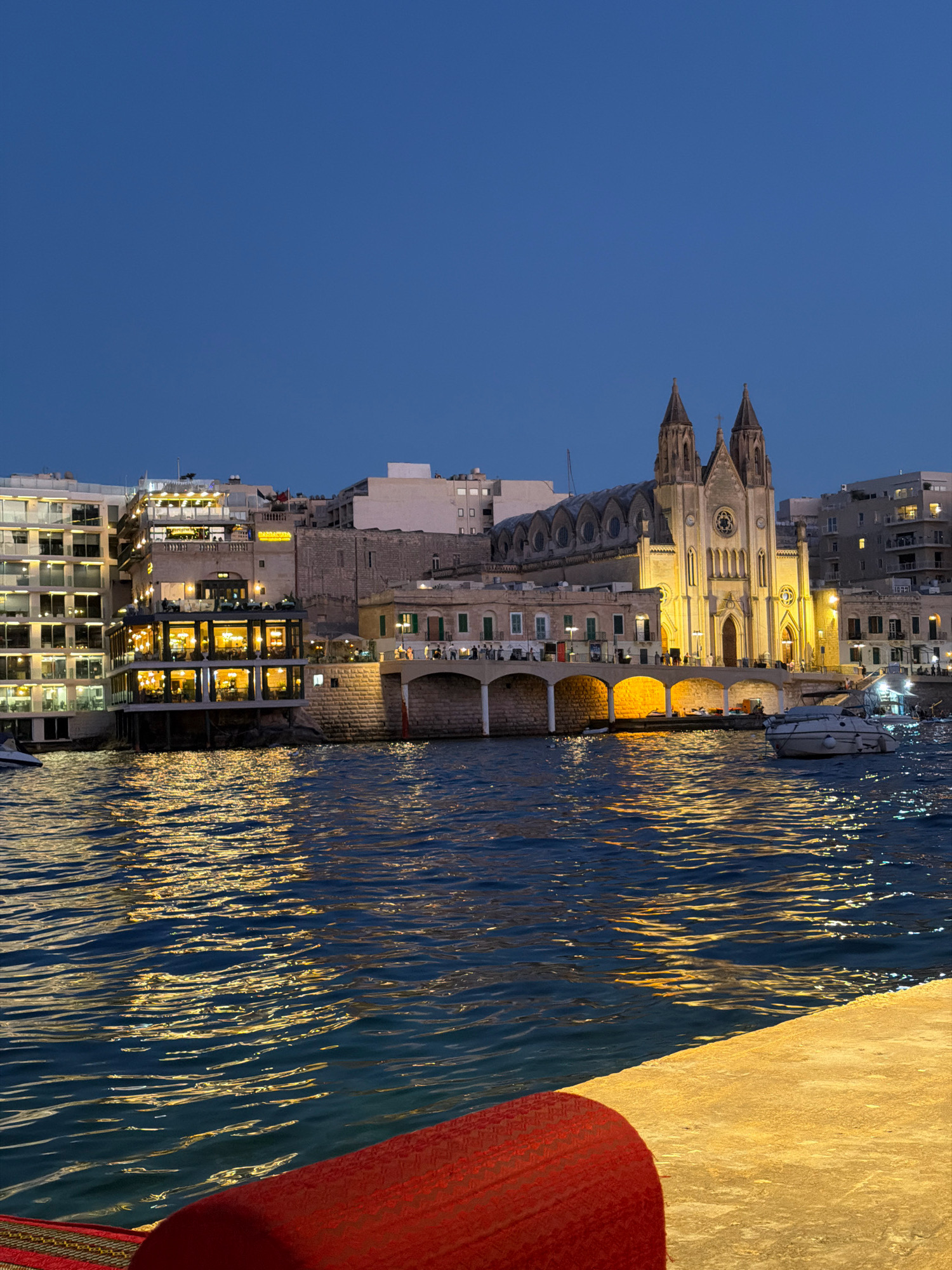 The image depicts a waterfront view of a European city at dusk, specifically San Ġwann, Malta. Dominating the skyline is a large, Gothic-style church with two prominent spires, illuminated by warm yellow lights. Adjacent to the church, a multi-story building with numerous windows, also lit from within, extends along the waterfront. A stone bridge with arched openings connects this building to the church's vicinity. In the foreground, dark blue water reflects the golden lights of the buildings, creating a shimmering effect on the surface. A portion of a red cushioned seating area is visible in the bottom right corner of the frame, suggesting a viewpoint from a boat or a waterside establishment. Several boats are moored in the water, and a few individuals can be seen walking along the illuminated waterfront promenade. The sky above is a deep, clear blue, characteristic of twilight.