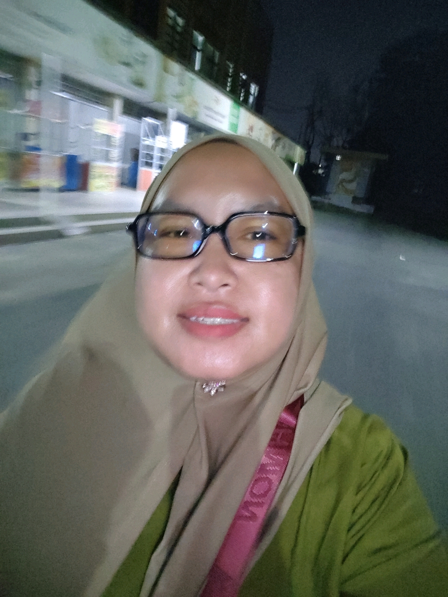In Batam, Indonesia, a woman wearing a tan hijab and green shirt is captured in a selfie at night. She has on black-rimmed glasses and a pink strap across her chest. The background shows a blurred street scene with a building on the left and a dark, cloudy sky on the right.
