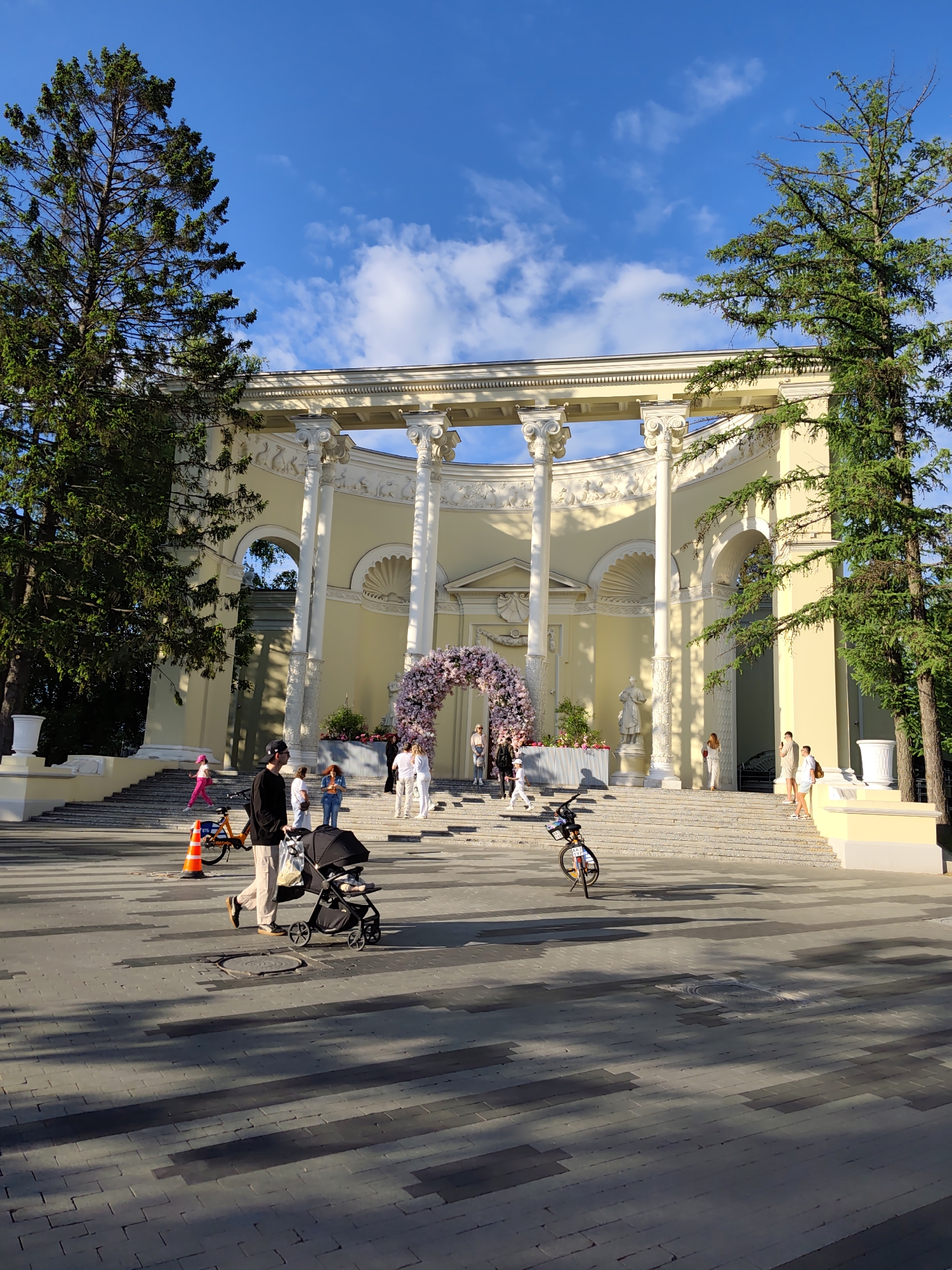 The image shows a daytime scene in a public park in Moscow, Russia. The dominant feature is a large, neoclassical-style pavilion with a colonnaded facade. The pavilion is light yellow and adorned with architectural details. A floral archway decorates the central steps leading up to the pavilion.
Several people are present in the scene. A man is walking across the foreground, pushing a stroller. A bicycle is nearby. Several other individuals are walking on the steps of the pavilion or standing near the entrance.
The surrounding environment includes tall trees on either side of the pavilion, framing the structure. The ground is paved with gray bricks. The sky is blue with scattered white clouds. A traffic cone is visible near the bicycle.