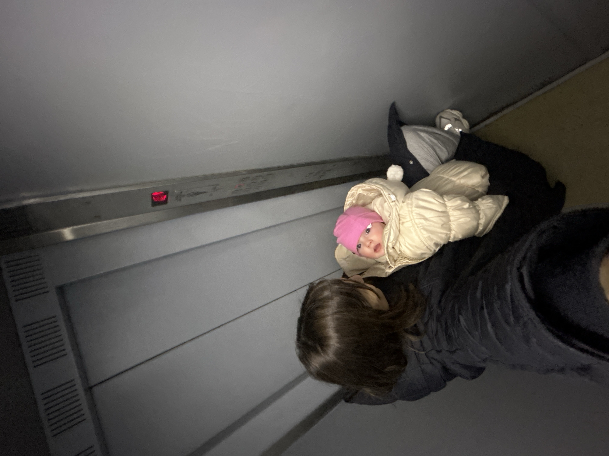 The image shows a person holding a baby, possibly inside an elevator. The person is wearing a dark jacket, and the baby is bundled in a light-colored snowsuit with a pink hat. An elevator panel is visible, suggesting the location is likely a building with multiple floors. It is difficult to discern any evidence to pinpoint this location as Moscow, Russia.