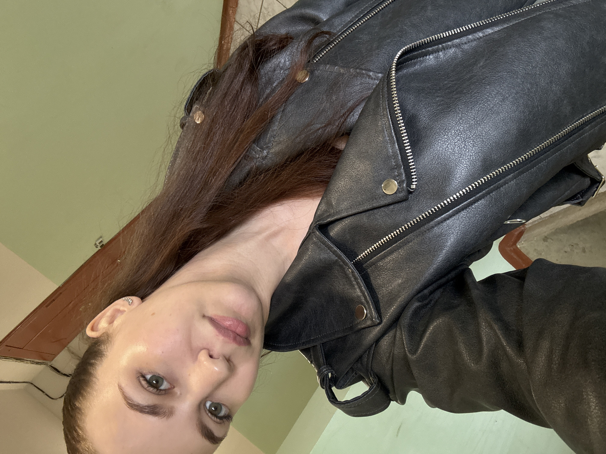 The image, oriented sideways, displays a young woman posing for a close-up photograph indoors. Her head is positioned on the left side of the frame, tilted slightly, and her body extends towards the right.
The primary subject is a woman with long, straight dark brown hair that flows over her shoulders and black leather jacket. She has fair skin, light-colored eyes that appear greenish-grey, and dark, well-defined eyebrows. Her expression is neutral to slightly contemplative, with a hint of a soft, closed-mouth smile. She wears subtle makeup, including a light pink lip color. In her visible left ear, she has a small, round silver stud earring in the lobe and a stacked silver cuff earring on the upper helix.
She is dressed in a black leather biker-style jacket, which features a textured, slightly worn finish. The jacket is adorned with numerous prominent silver zippers and several metallic gold or brass snap buttons on the lapels and shoulders.
The background is predominantly a plain, light olive-green wall. In the upper part of the image (when viewed upright), a dark brown wooden element, possibly a door frame or a piece of furniture, is partially visible. Some dark wires are also seen running along or near this wooden structure. The lighting suggests an indoor setting.
No text is visible in the image.