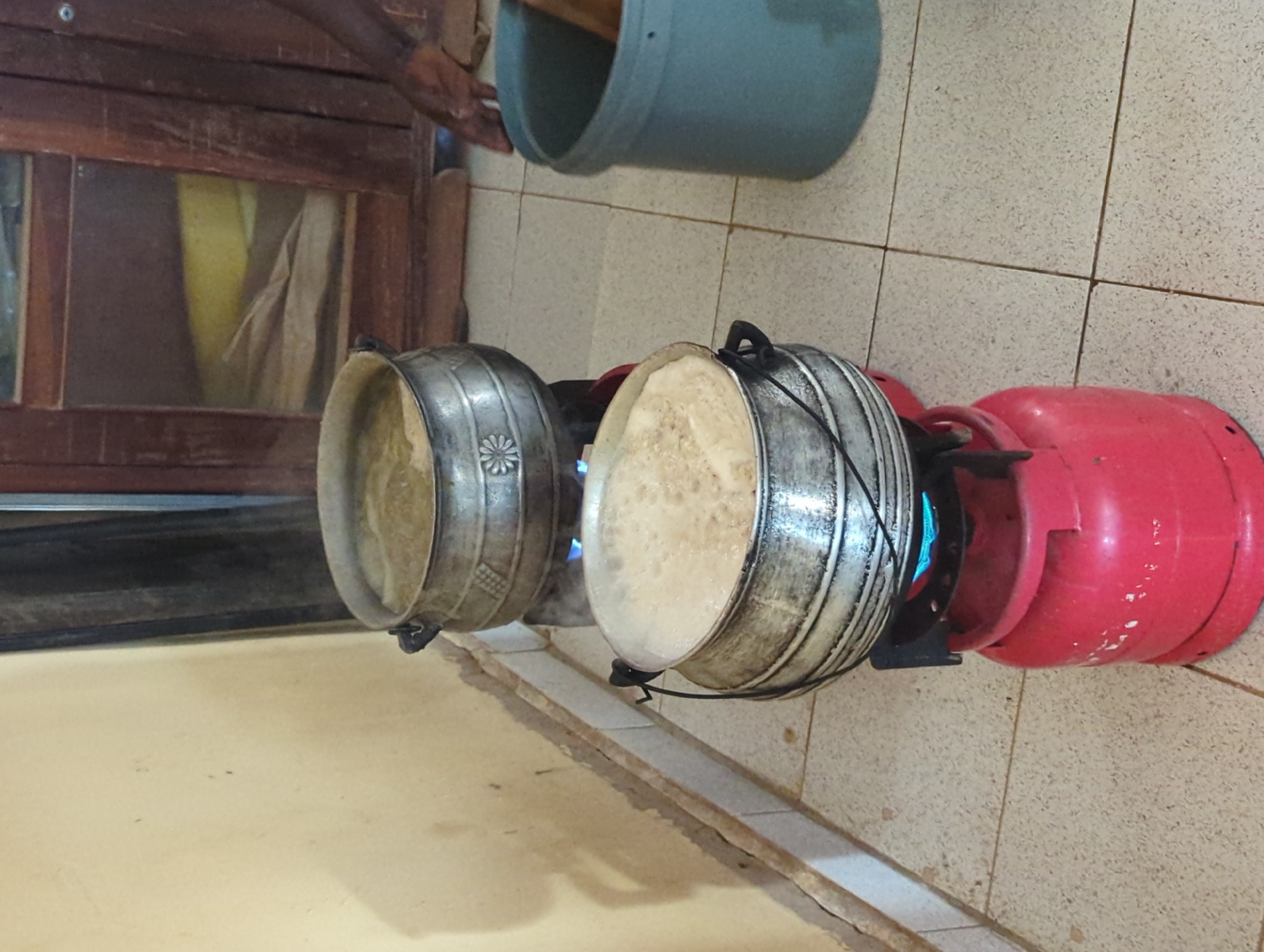 Two metallic pots containing a light-colored substance that appears to be boiling are set on separate portable gas stoves with pink propane tanks on a tiled floor in a room with a wooden-framed window. One hand is visible near a gray bucket, and the details suggest a kitchen-like environment. The location is not identifiable, and further information is needed to provide the city and region.