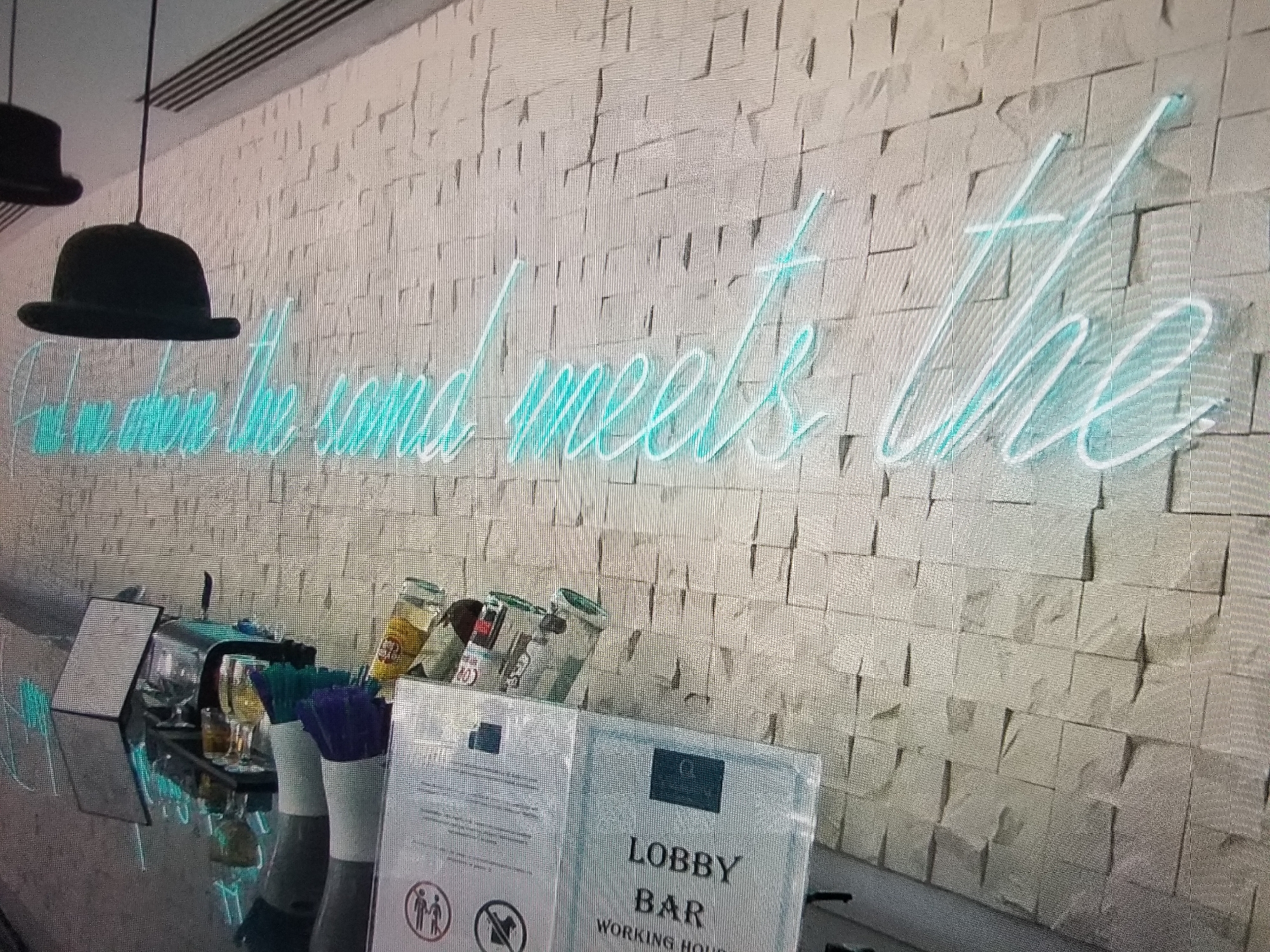 The image depicts an interior scene with a wall textured with light-colored rectangular blocks. A neon sign in a light blue color is mounted on the wall. The text on the neon sign reads "when the sand meets the". Two bowler hats are visible hanging from the ceiling on the left side of the image. In the foreground, a bar counter is present with various items including glasses, bottles, and what appear to be cocktail stirrers in holders. A sign that says "LOBBY BAR WORKING HOURS" is also visible on the counter. The overall lighting suggests an indoor environment, possibly a bar or lounge area. The location is specified as Tiraspol, Moldova. There are no people visible in the image.