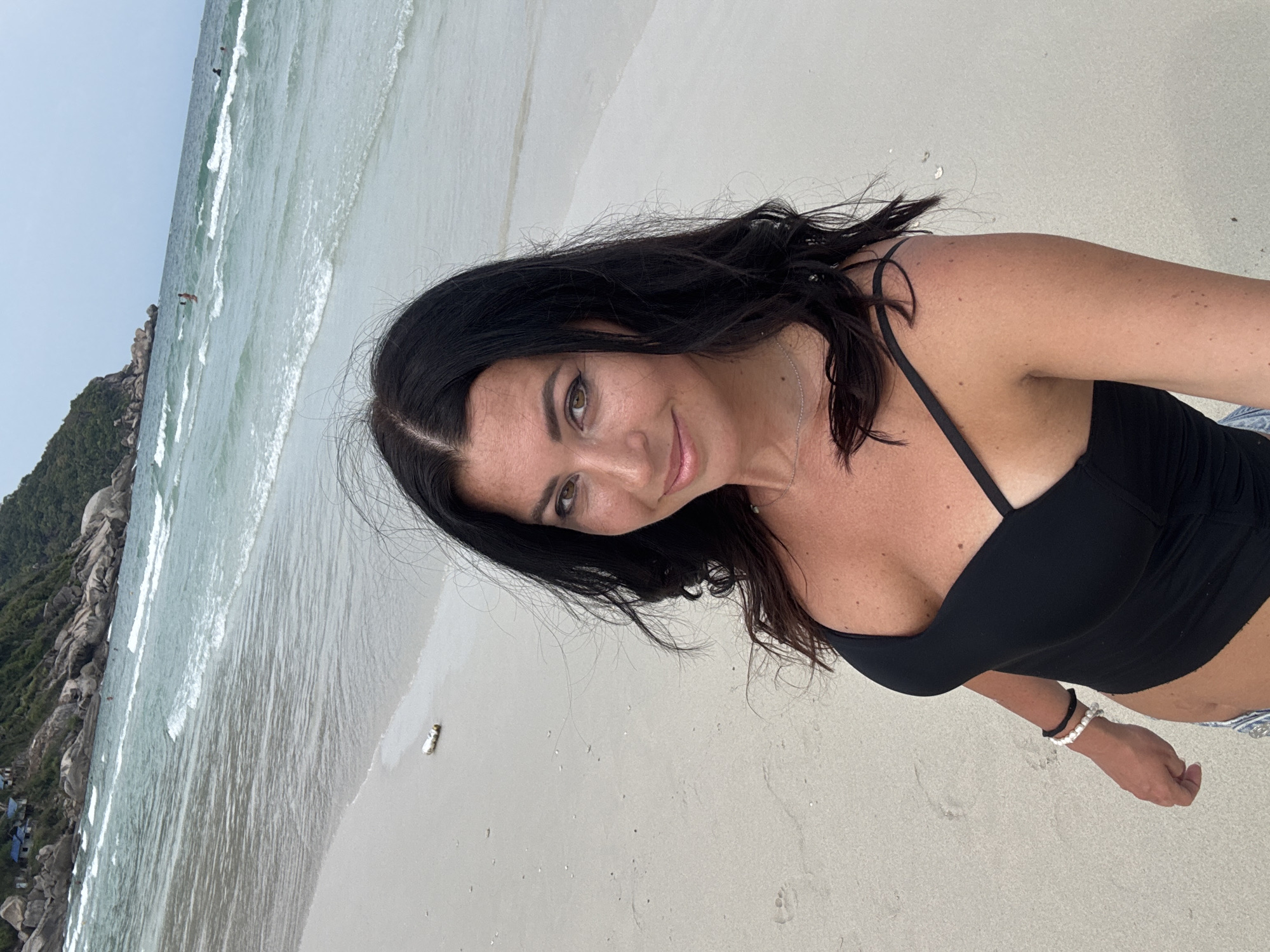 A woman with dark hair and a black top is on a sandy beach in Ban Haad Rin, Thailand. The beach has light-colored sand and is next to a body of water with small waves. In the distance to the left, there is a rocky, green landscape. The sky is overcast.