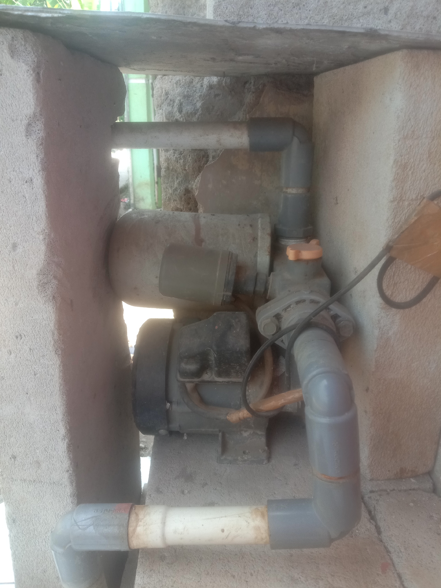 A close-up, slightly blurry, horizontal outdoor shot shows a water pump system constructed from PVC pipes and a motor, partially embedded within rough concrete blocks in Cianjur, Indonesia. The pump, a dark motor unit with a cylindrical component above it, is connected to various gray PVC pipes forming elbows and straight sections. An orange cap is visible on the pump's housing. The concrete structure appears unfinished, with some gaps and rough textures. The background is out of focus, showing a hint of greenery and a bright sky. The lighting suggests it is daytime.