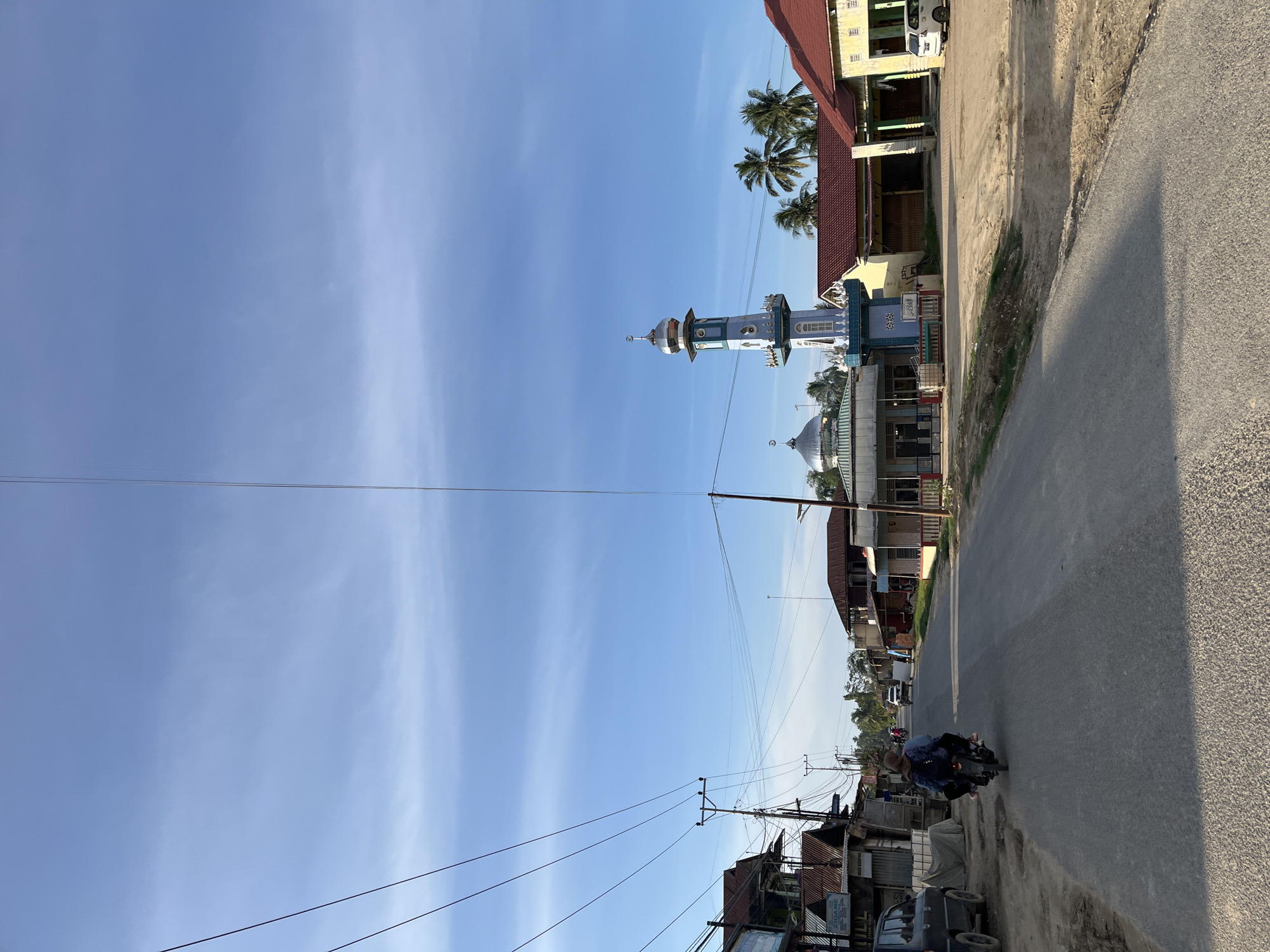 This media file captures a street scene in Alurcucur, Indonesia, under a bright, clear blue sky with some wispy white clouds. The photograph is oriented vertically, with the road running diagonally from the bottom right towards the top left.
The scene features a street lined with buildings on the left side, including a blue and white mosque with a tall, slender minaret topped with a silver dome. Palm trees are visible behind some of the buildings. The buildings appear to be a mix of residential and commercial structures, with some showing signs of wear. A paved road occupies the right side of the frame, with a rougher shoulder on the far right.
A person is riding a motorcycle on the road, heading away from the camera. The rider is wearing a blue shirt. The time of day appears to be late morning or early afternoon, judging by the shadows cast by the buildings and the bright sunlight. The weather seems pleasant and sunny.
Several utility poles with overhead wires are prominent in the scene, indicating infrastructure. Some small structures or makeshift buildings are also visible along the roadside. No significant text is legible in the image, although a sign with the word "ZODANE" is partially visible on a building. The overall impression is of a typical day in a rural or semi-urban Indonesian community.