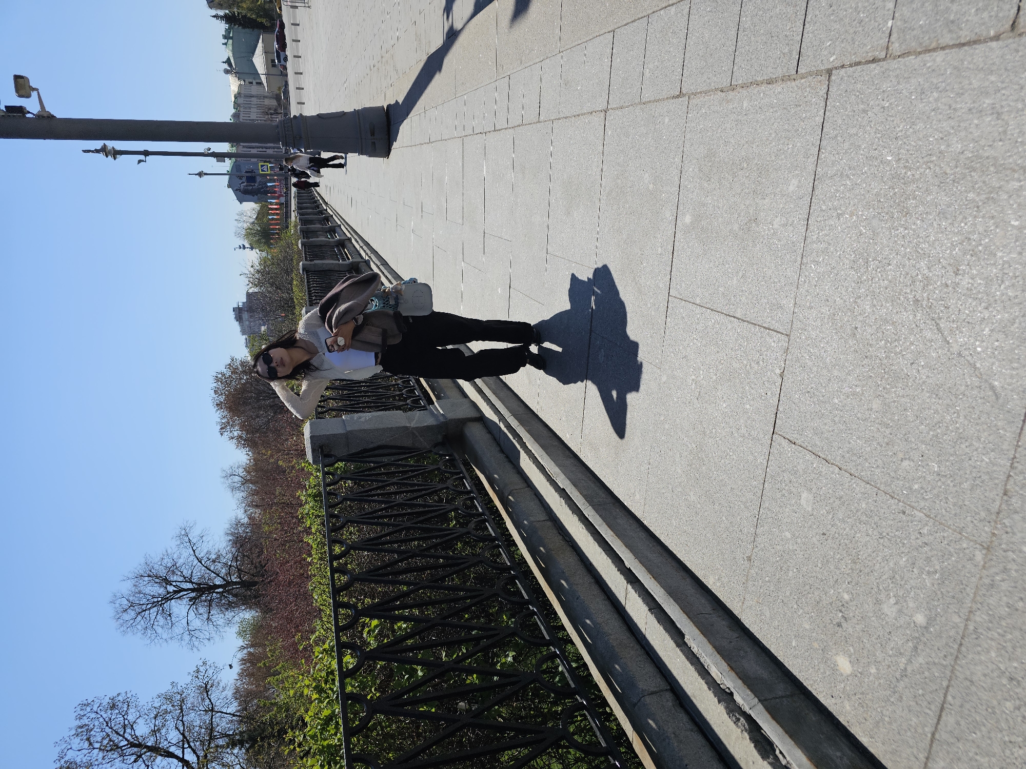 A woman stands on a wide pedestrian walkway in Moscow, Russia. The walkway is paved with large, light-gray rectangular stones. A black metal railing runs along the walkway's edge, separating it from a lower area with trees and other vegetation. The woman is wearing sunglasses, a light-colored top, dark pants, and is carrying a bag. Her shadow stretches onto the walkway beside her. In the background, across the walkway, there are historical buildings and lampposts. The sky is blue and clear.