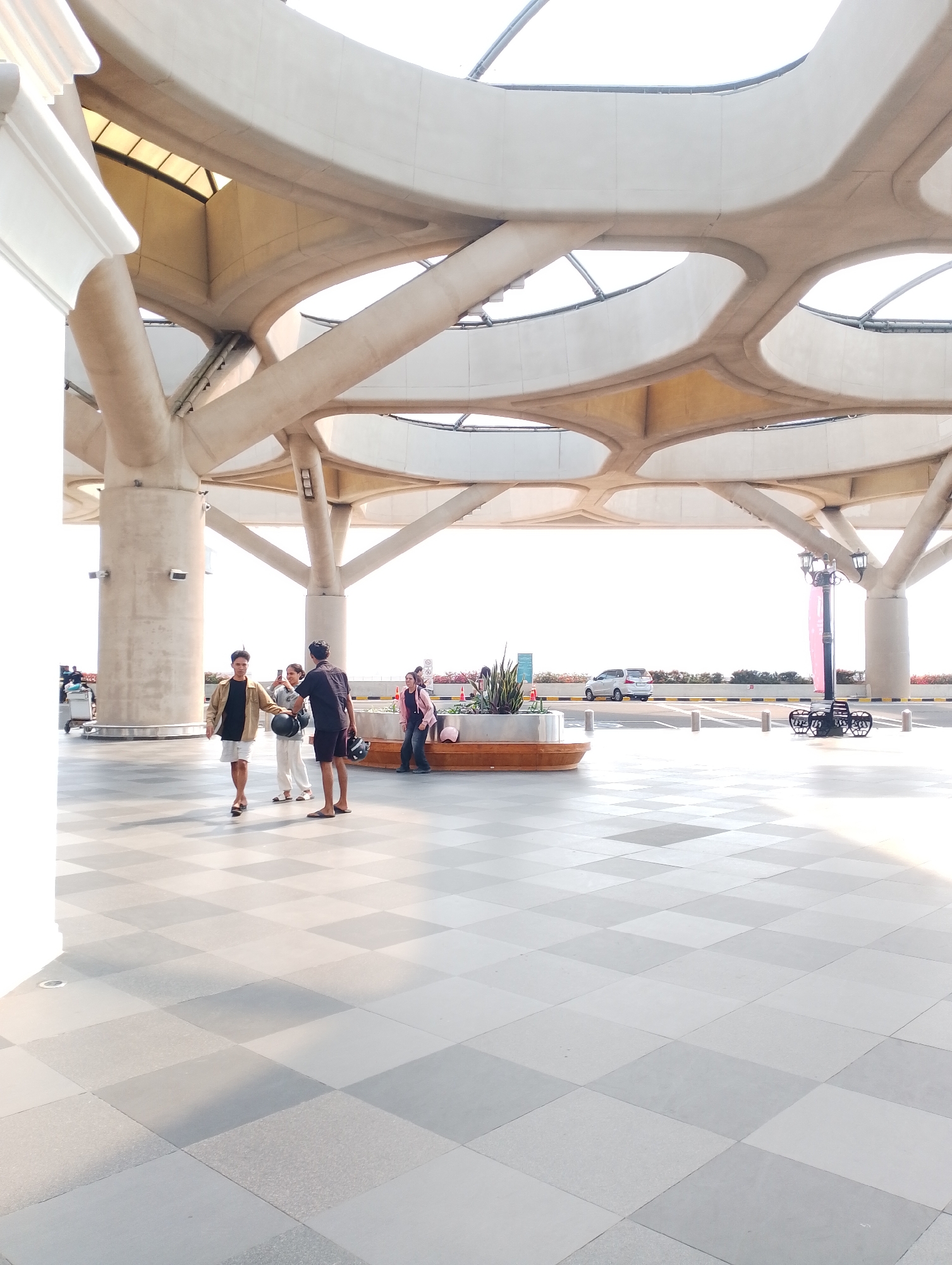 The image depicts the exterior of a modern building with a distinctive, organic architectural design featuring large, curved concrete beams and columns that form an open canopy. The ground is paved with a checkerboard pattern of light and dark gray tiles. Sunlight streams in from the right, casting shadows.
In the foreground, a group of three people stands on the tiled area. A young man in a tan jacket and white shorts walks towards the camera, looking to his right. To his left, a person in a white top and pants stands facing him, holding a dark circular object. A third person, wearing a dark short-sleeved shirt and shorts, stands to the right of the person in white, facing the first man and holding a helmet.
Further back, a white car is visible on a road beyond a yellow and black striped barrier. A black metal bench with an ornate design is situated near a lamppost. In the middle ground, there is a wooden bench surrounded by plants. Another person is visible to the far right, wearing a pink jacket and jeans, and appears to be interacting with someone out of frame. The overall environment suggests an outdoor public space, potentially an airport or transportation hub, given the presence of luggage trolleys on the left and the open, airy design. The location is identified as Palihan, Indonesia.